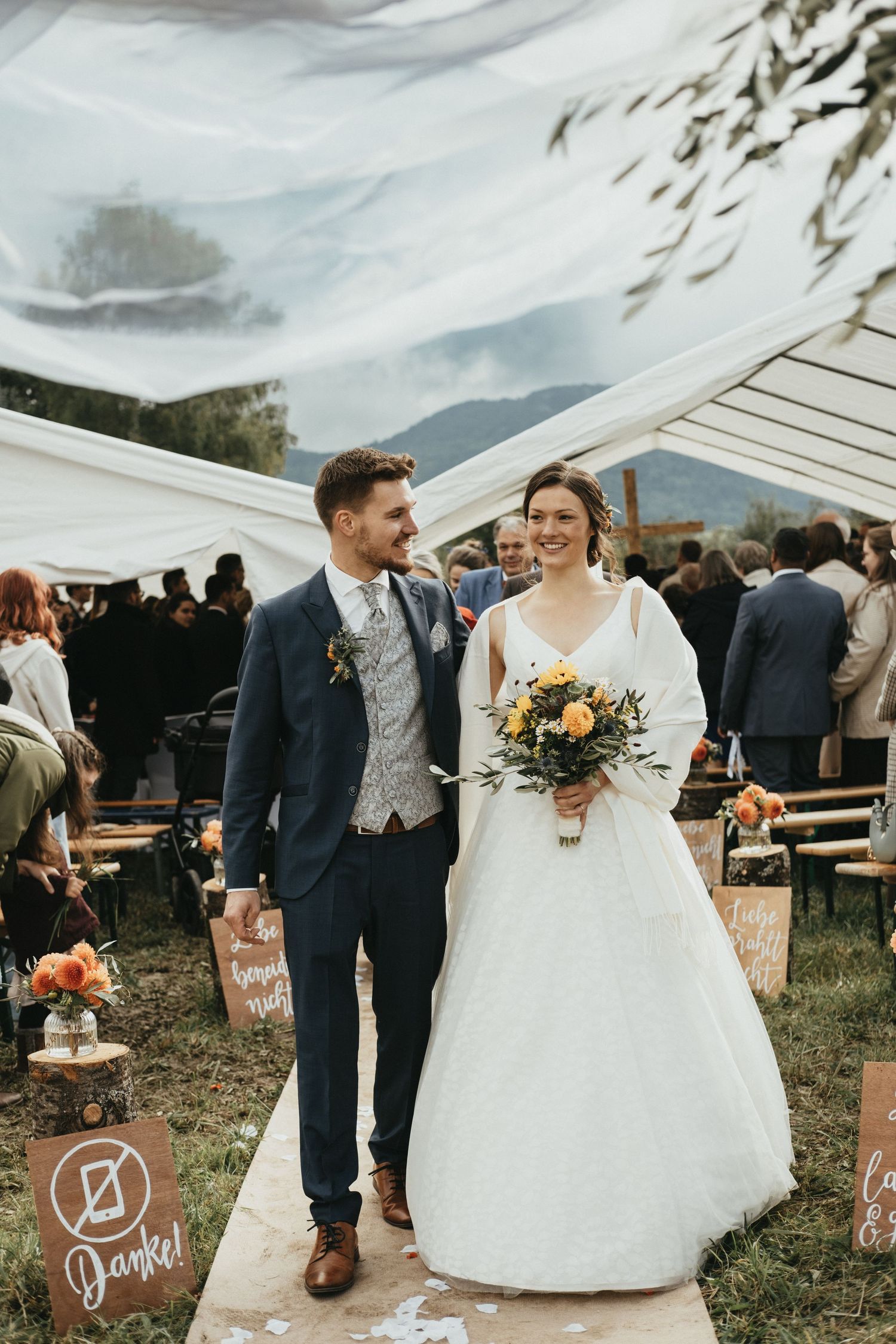 A couple celebrates their rustic outdoor wedding with guests in a field under a cloudy sky.
