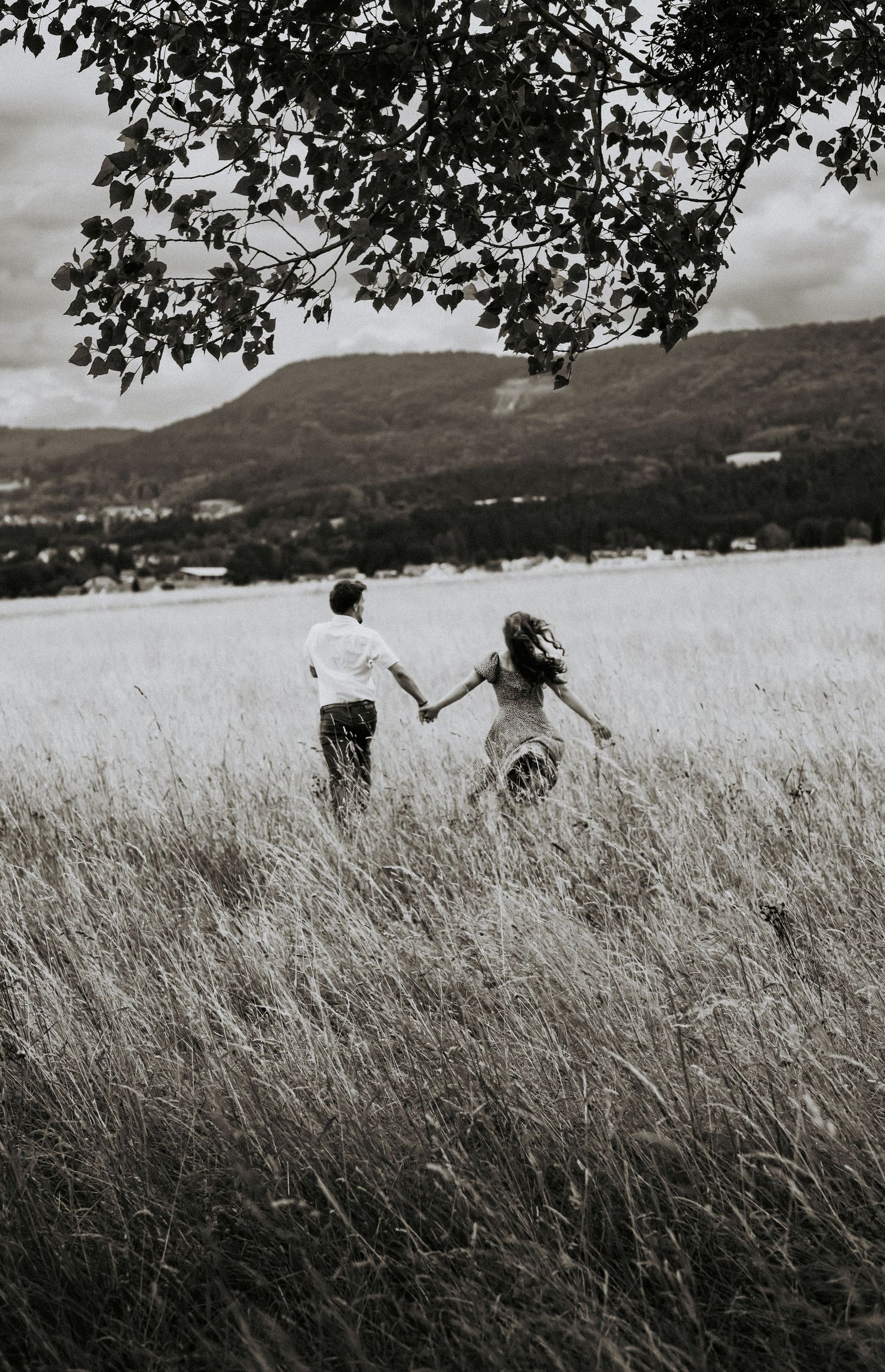A couple runs hand in hand through a tall grass meadow with mountains in the background in black and white.
