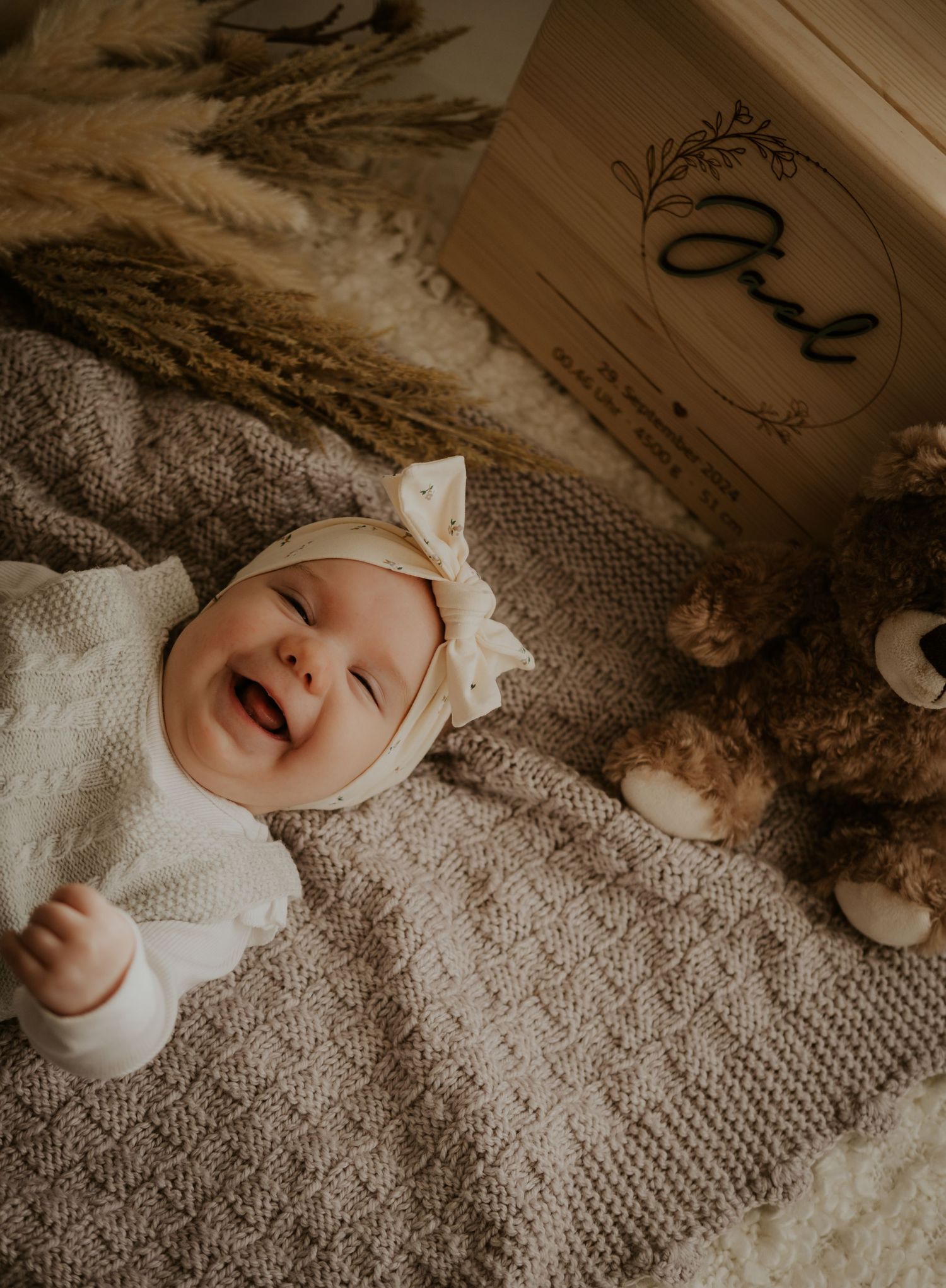 A baby lying on a cream knit blanket smiles and laughs while wearing a white bow headband.
