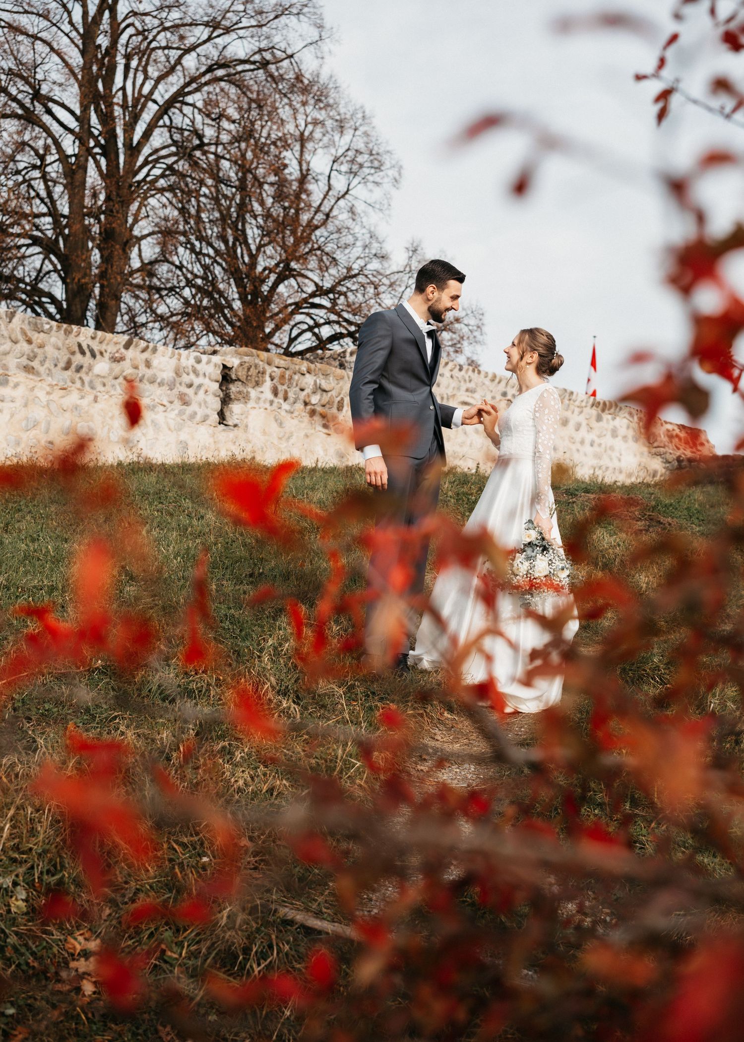 Newlywed couple shares romantic moment on stone wall with red autumn foliage in foreground.