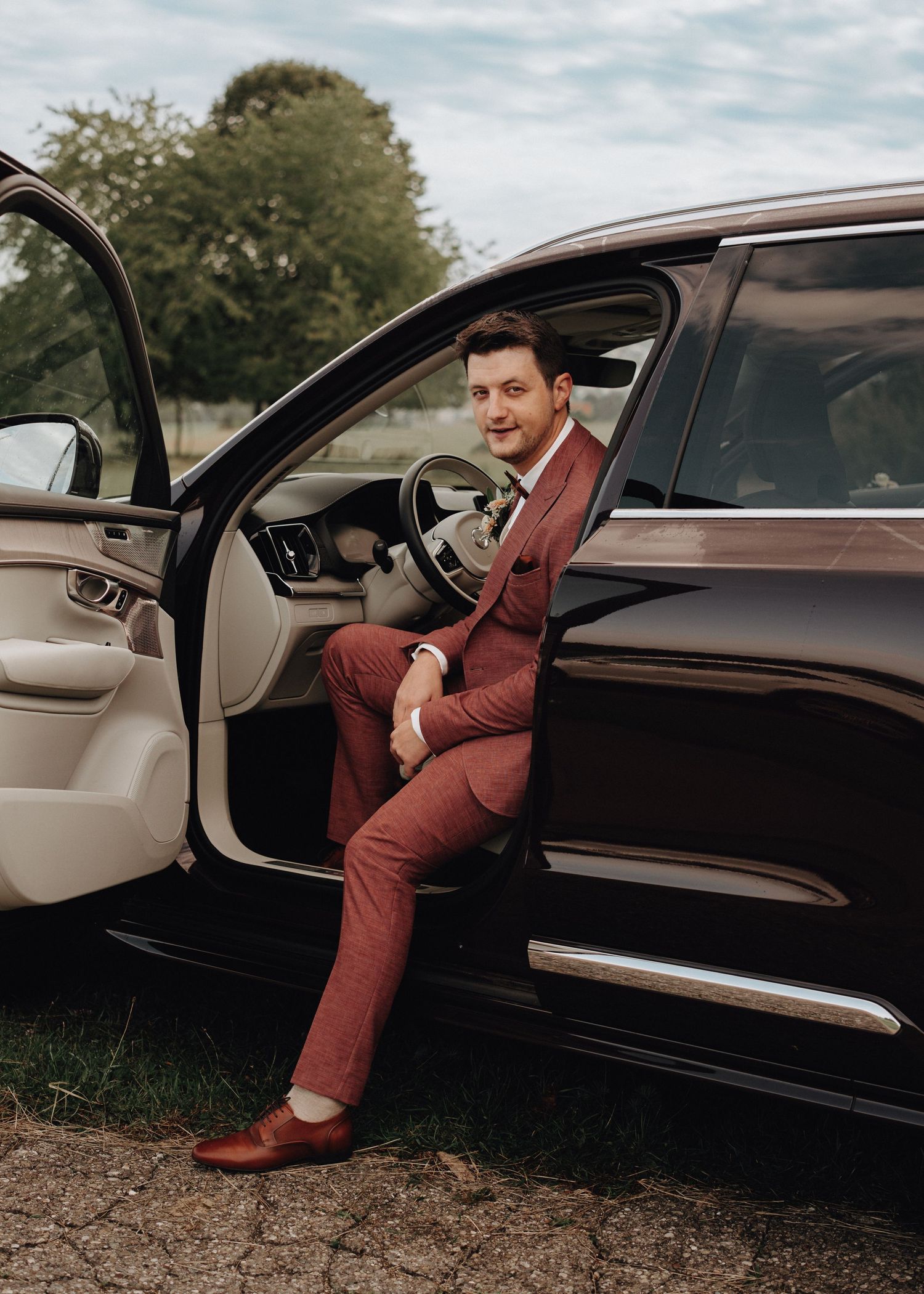 A person in a burgundy suit sits in a luxury car with beige leather interior.