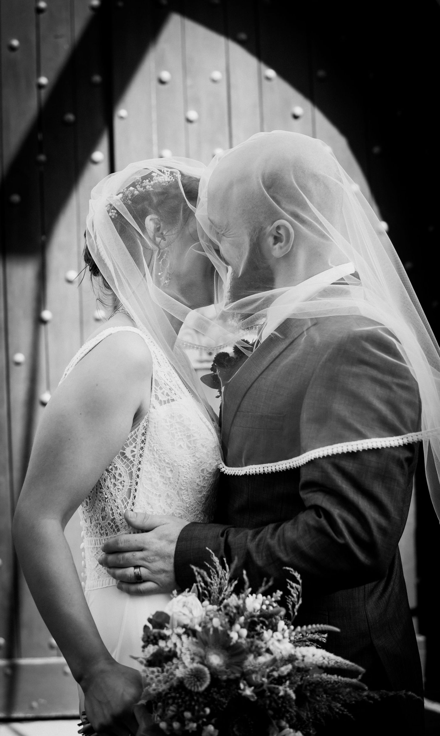 A romantic black and white wedding photograph showing a couple kissing under a delicate veil with a bouquet in hand.