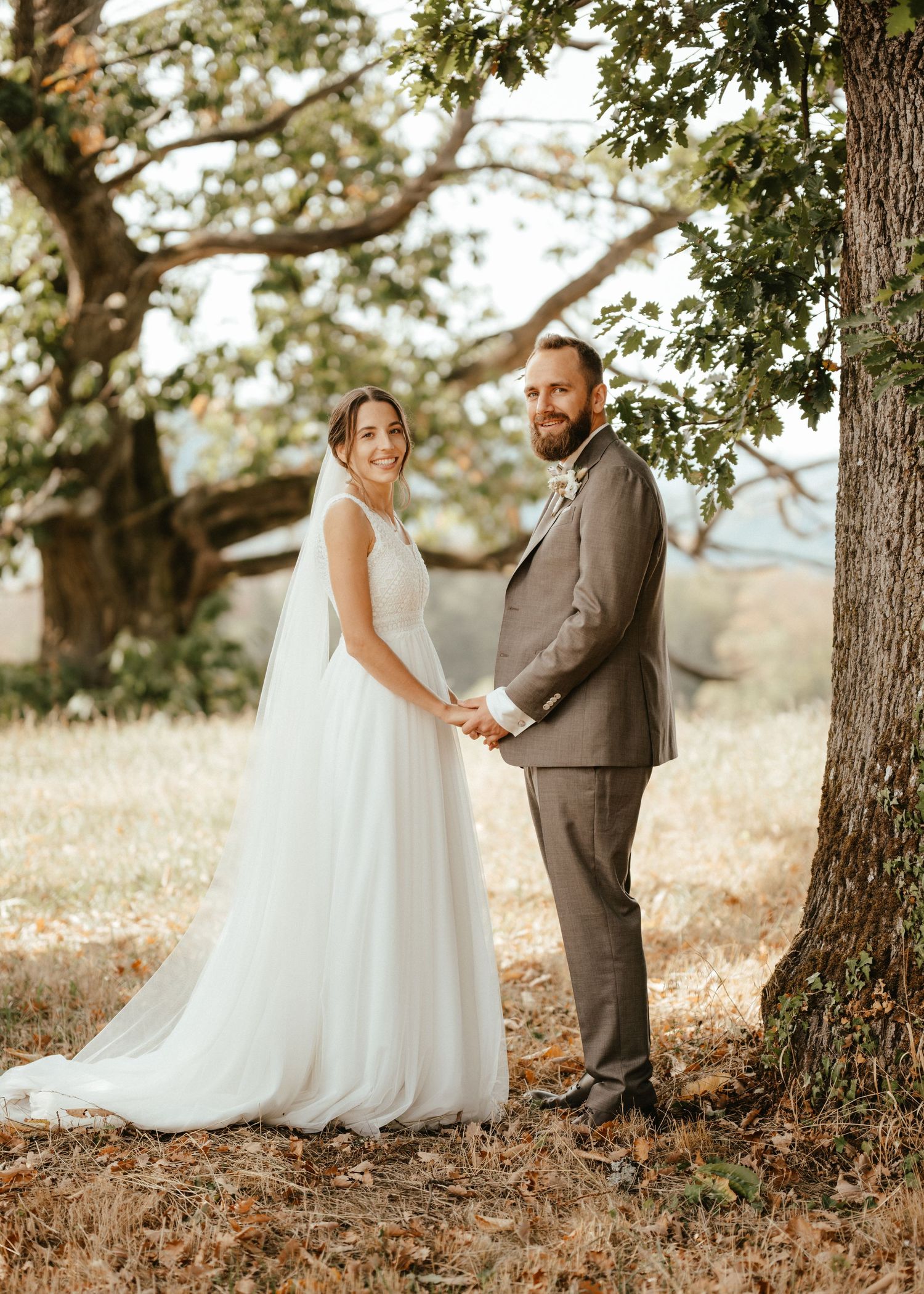 A bride in a white gown and groom in a tan suit hold hands near a large tree at an outdoor wedding.