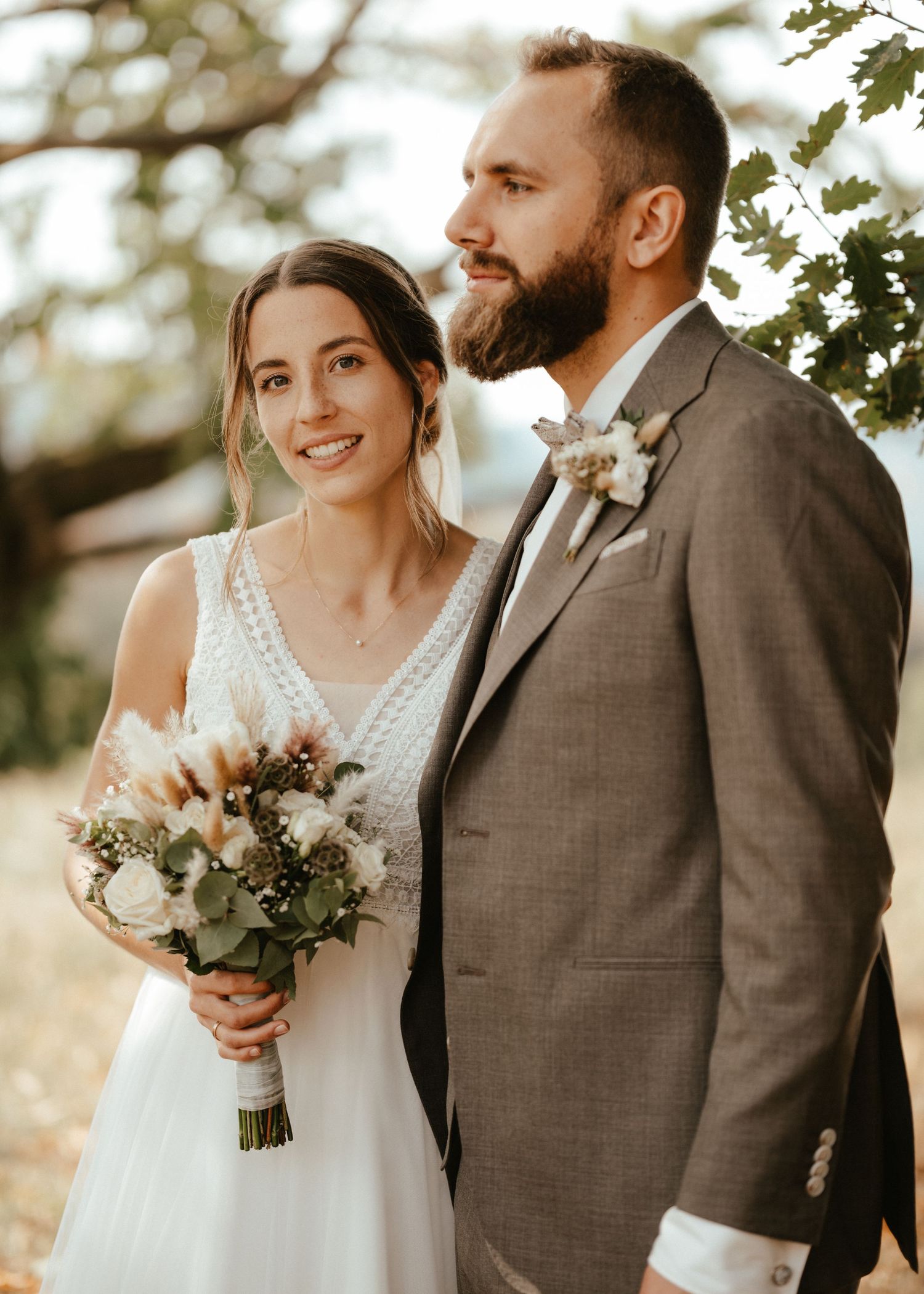 A smiling bride holding a bouquet stands beside a bearded groom in a gray suit with a boutonniere outdoors.
