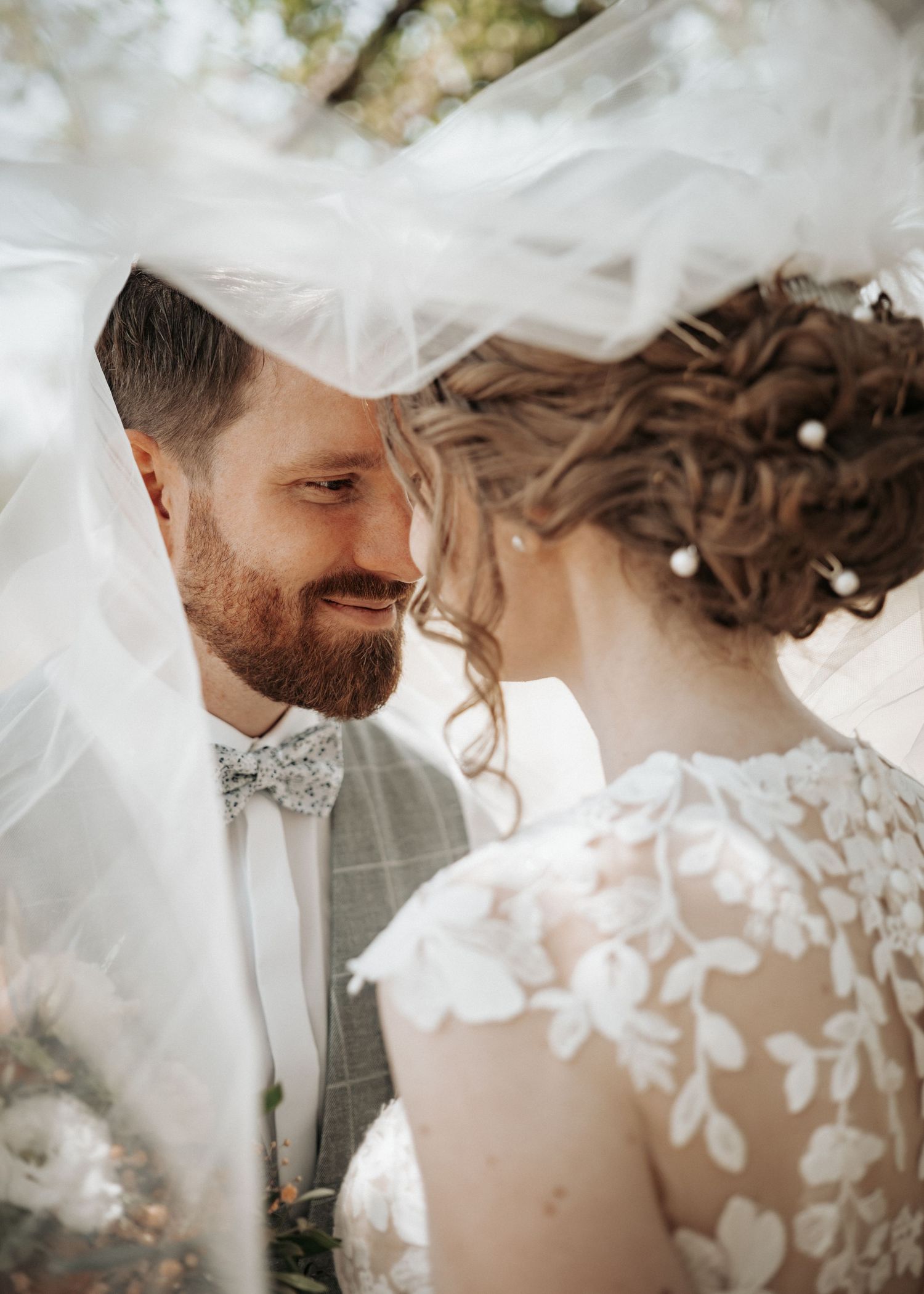 A smiling bearded groom and bride share an intimate moment under a flowing white wedding veil outdoors.