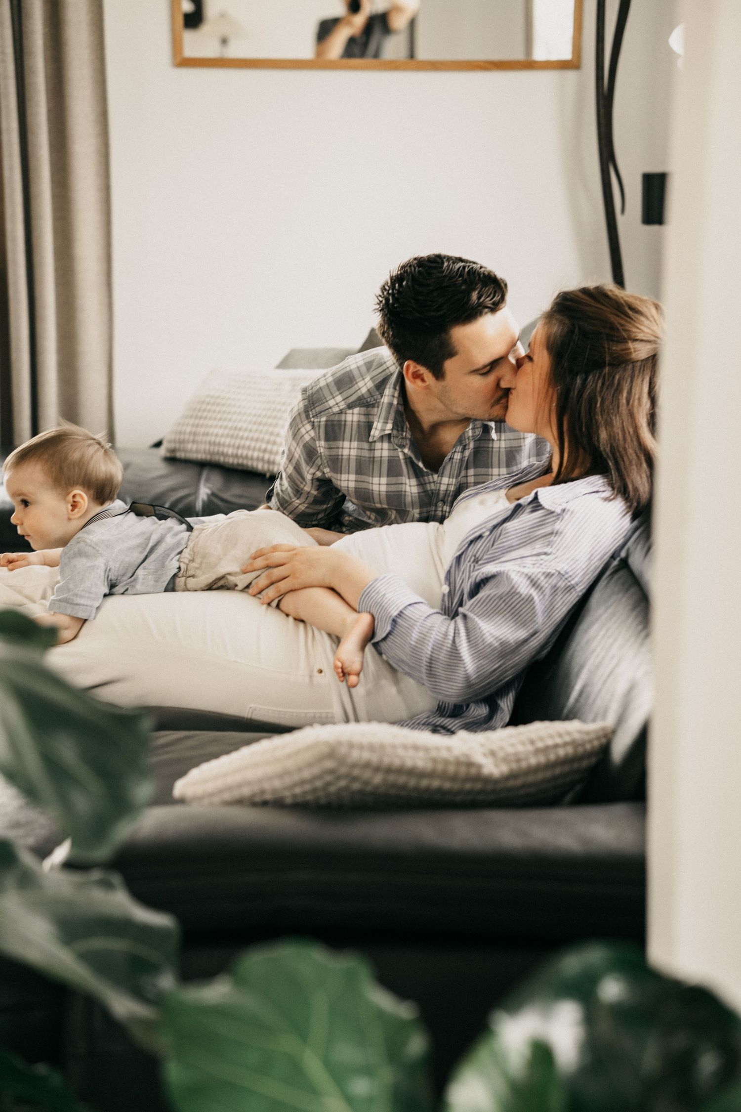 A couple relaxes together on a gray couch in a cozy living room with green plants visible in the foreground.