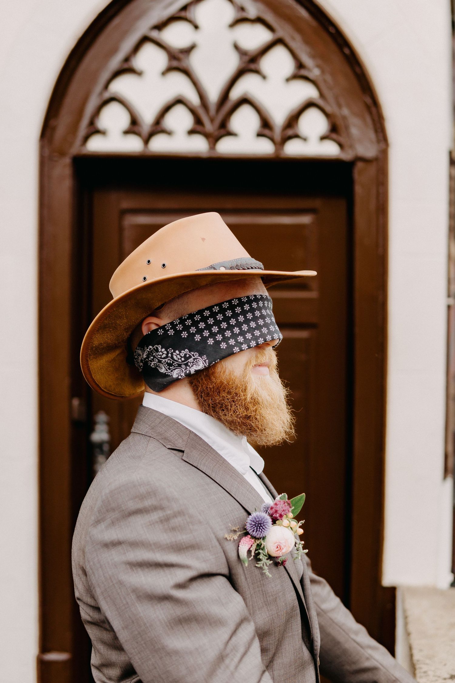 A person in a gray suit and cowboy hat with a boutonniere stands in front of ornate wooden doors.