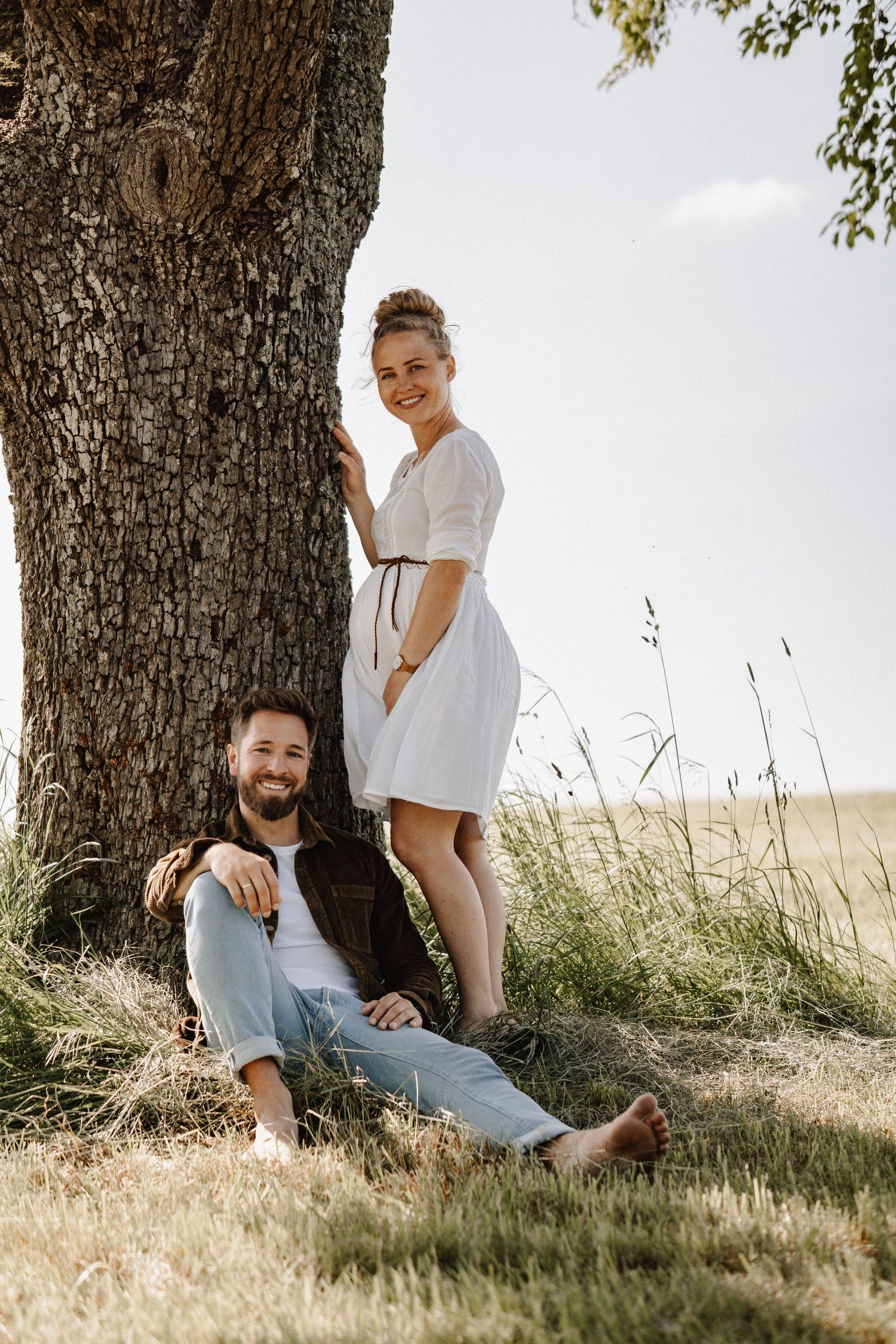 Couple relaxes together under a large tree in a sunny field with natural lighting and casual summer attire.