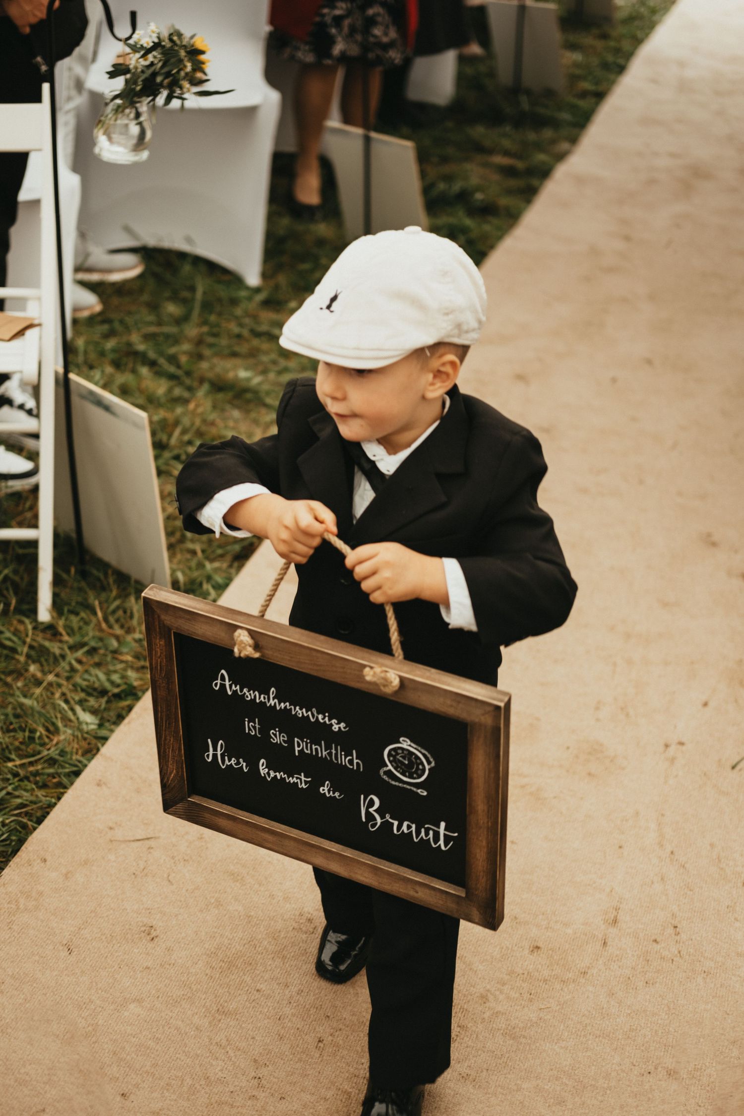 A young ring bearer in a black suit carries a rustic chalkboard sign down a wedding ceremony aisle.