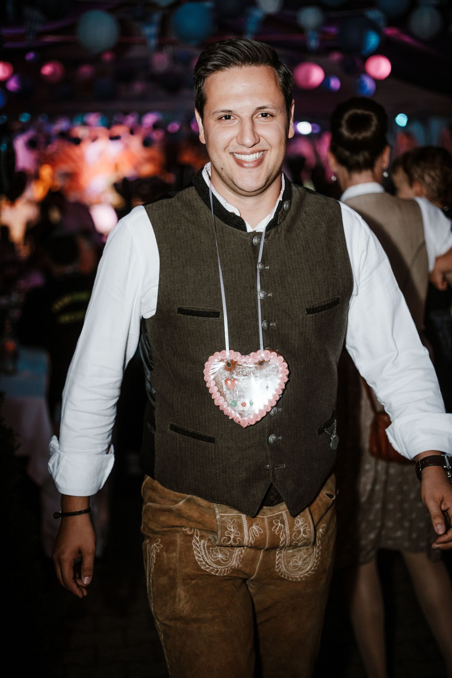 Person wearing traditional Bavarian lederhosen and white shirt dancing at a festive event with colorful lights in background.