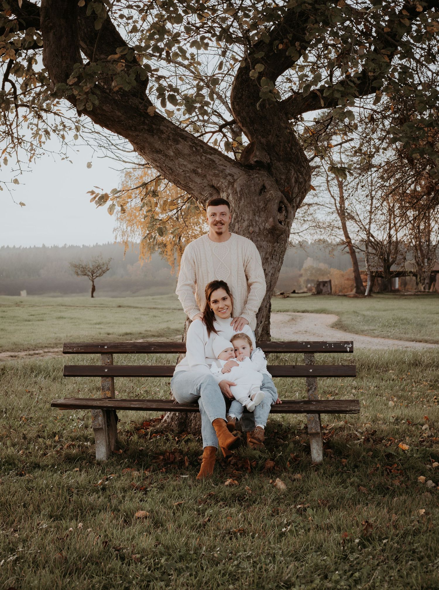 A family sits together on a wooden bench under a large tree during a golden autumn evening in a peaceful meadow.