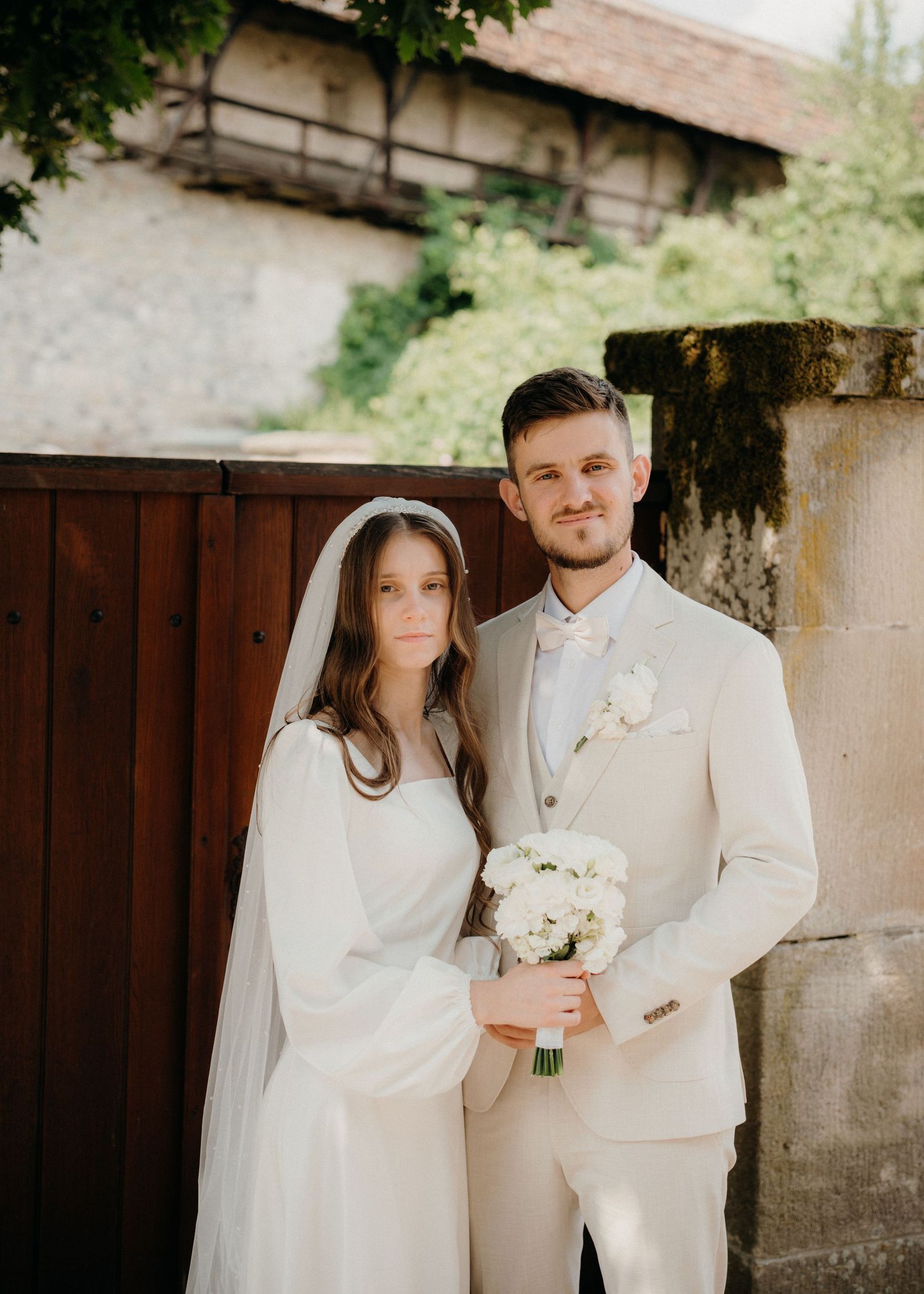 Bride and groom posing outdoors in elegant white wedding attire, holding a white floral bouquet near a rustic stone wall.