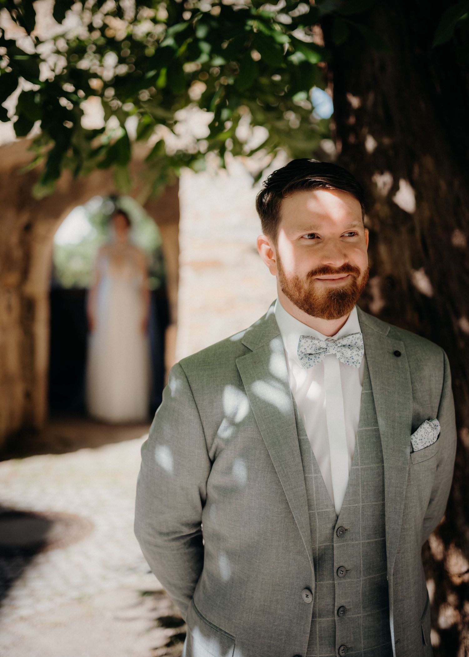 Groom in gray suit and bow tie smiling outdoors, bride visible in blurred background beneath tree.