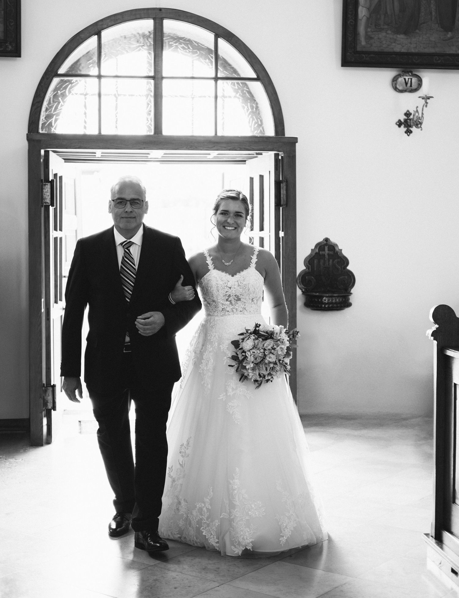 Black and white photo of a wedding processional walking through an arched doorway in a church hallway.