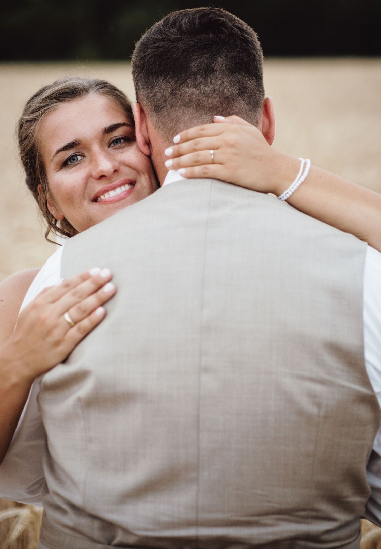 A couple embraces during a wedding photoshoot, with the bride smiling warmly while hugging her partner in a beige vest.