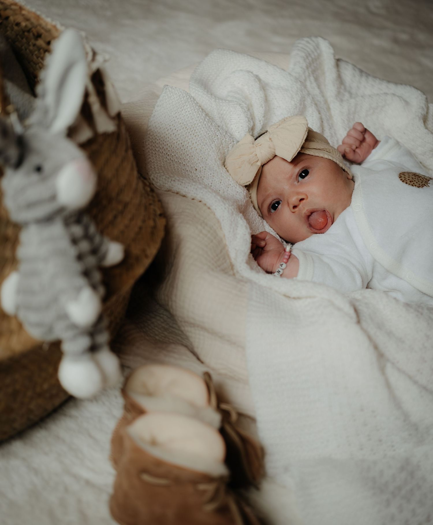 A newborn wearing white clothing rests on soft white bedding with plush bunny toys nearby.
