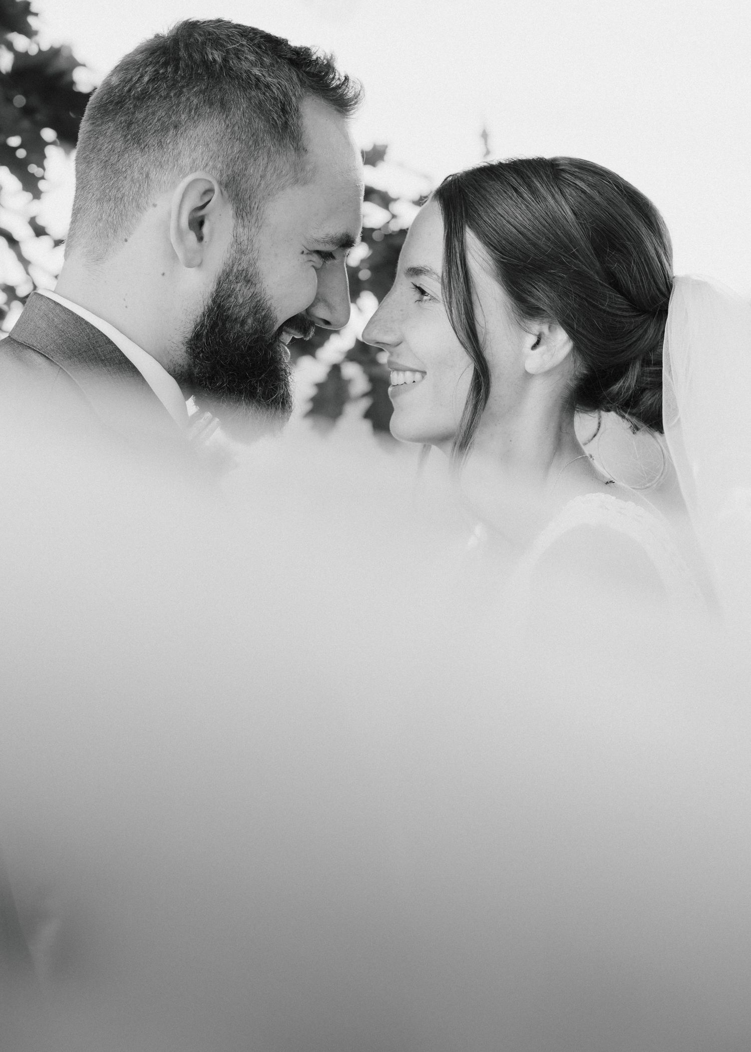 Black and white wedding photo of smiling bride and bearded groom gazing at each other outdoors.