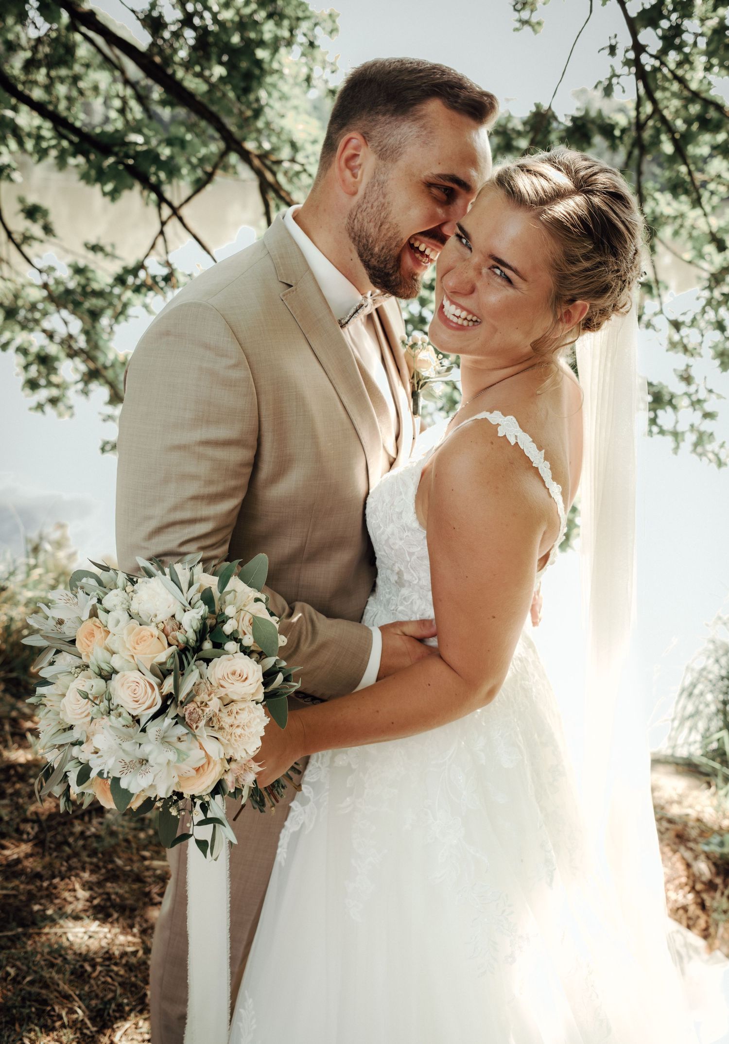 Newlyweds share joyful moments under tree branches with white floral bouquet.