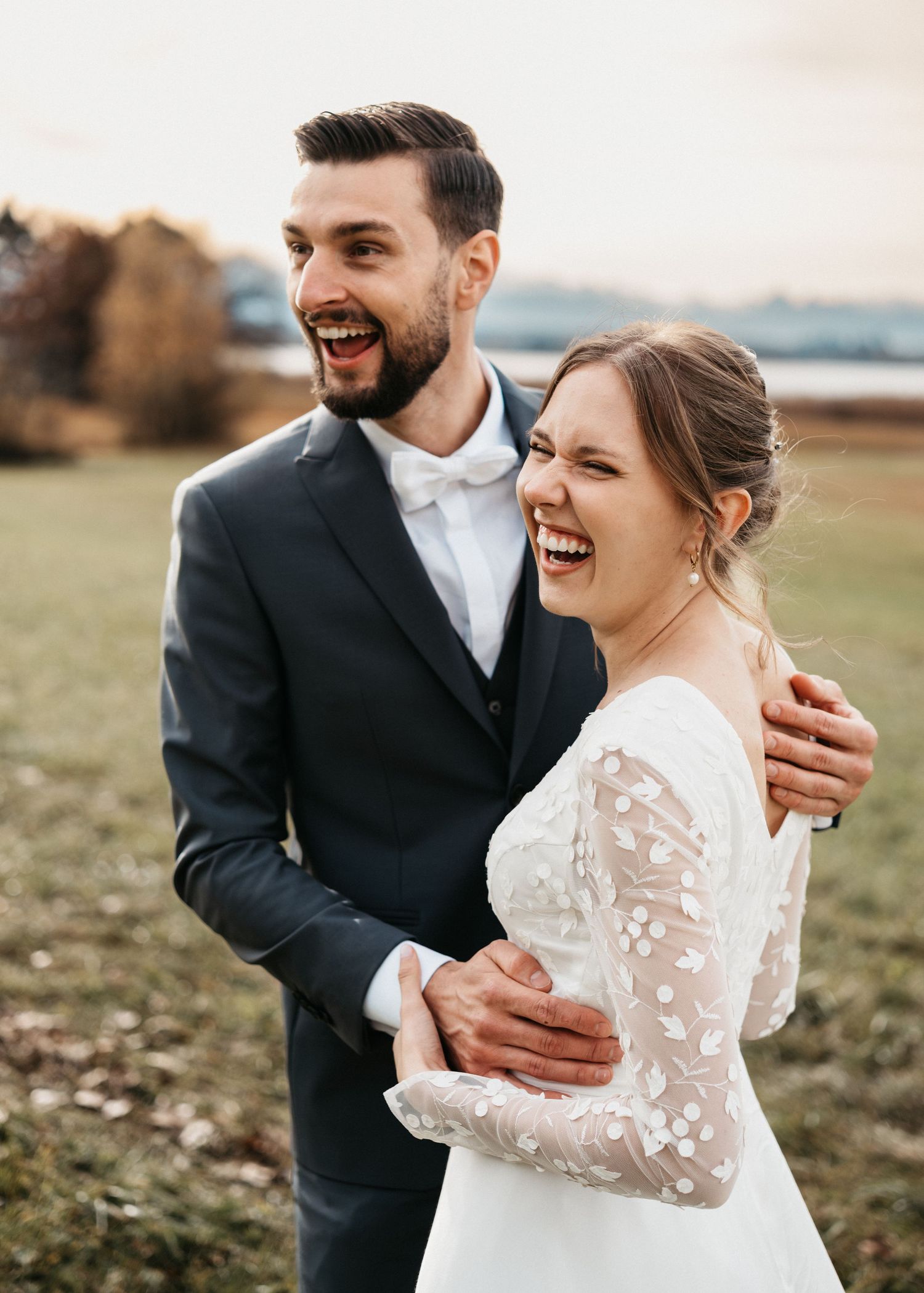A joyful couple laughs together in an outdoor autumn field during their wedding photoshoot.