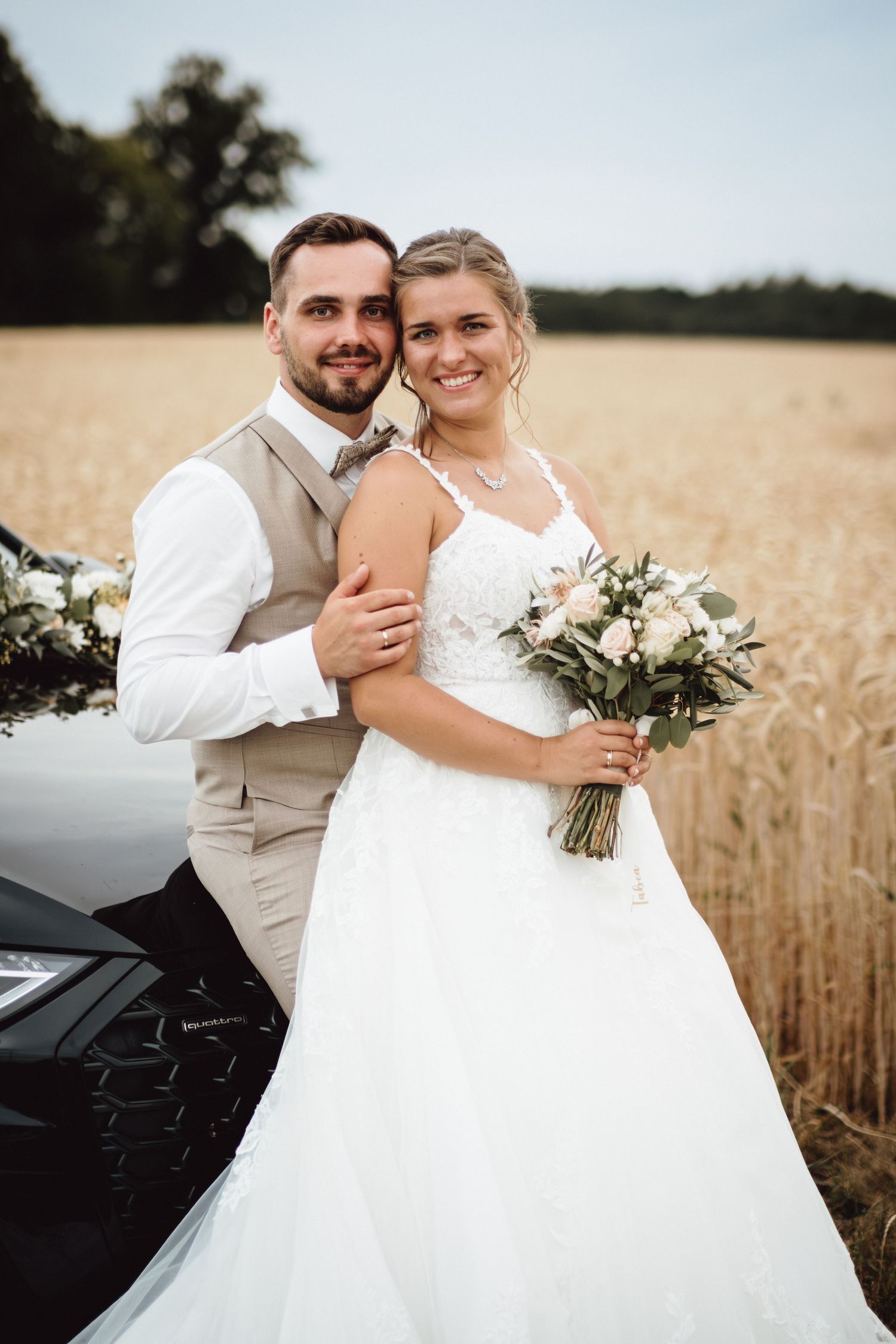 A romantic wedding photoshoot in a golden wheat field with a black car in the background.