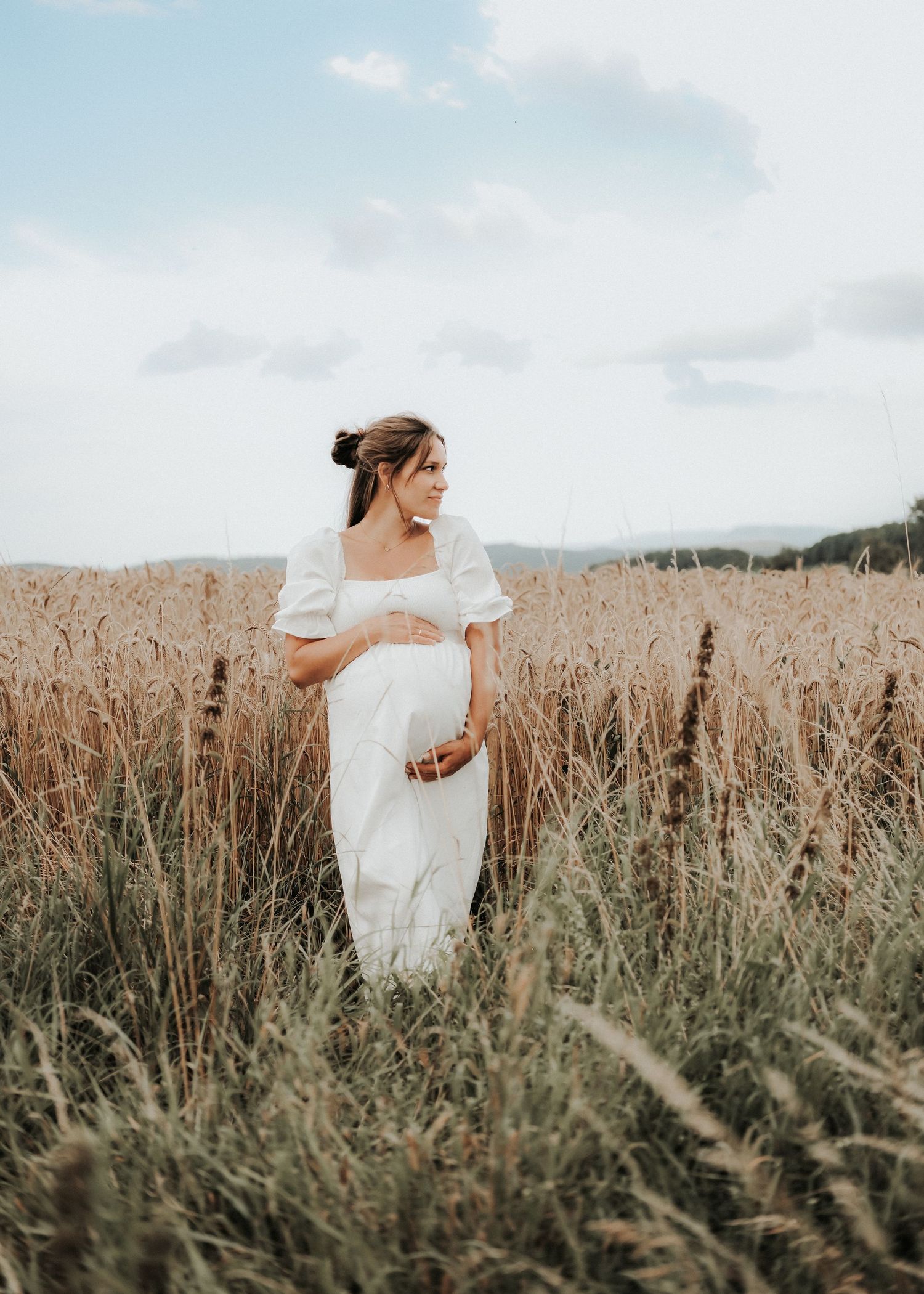 A bride in a flowing white dress walks through a golden wheat field on a cloudy day.