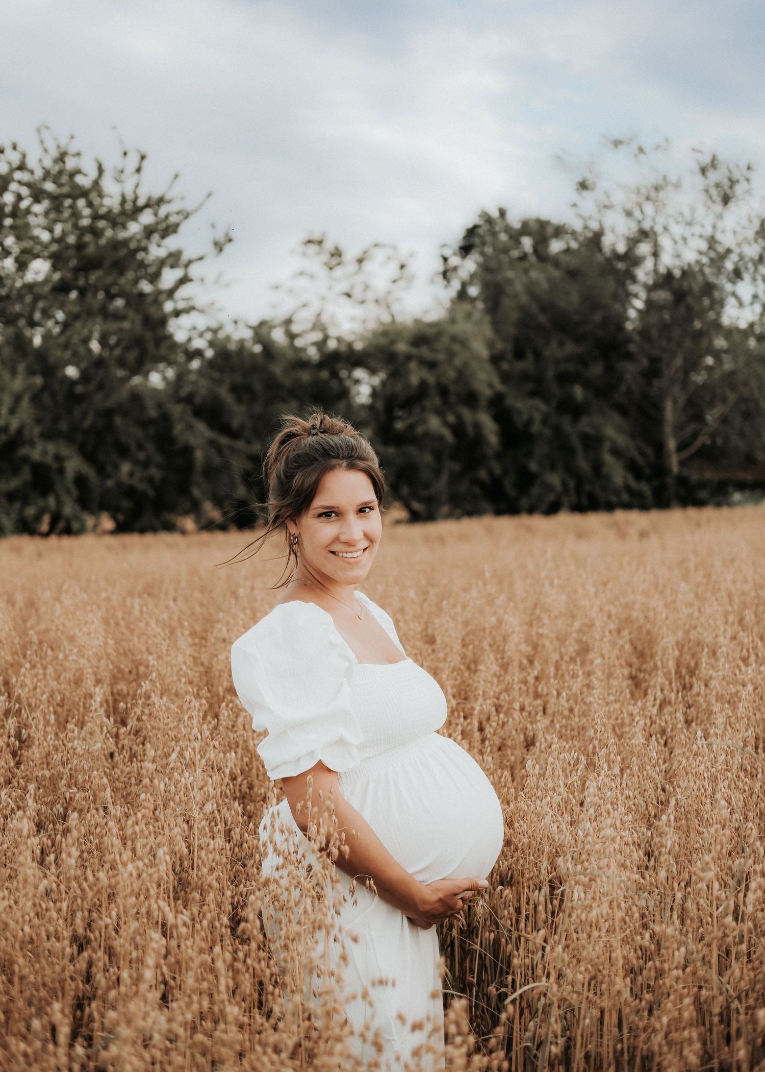 Expectant mother in white dress poses in golden wheat field during sunset maternity photoshoot.