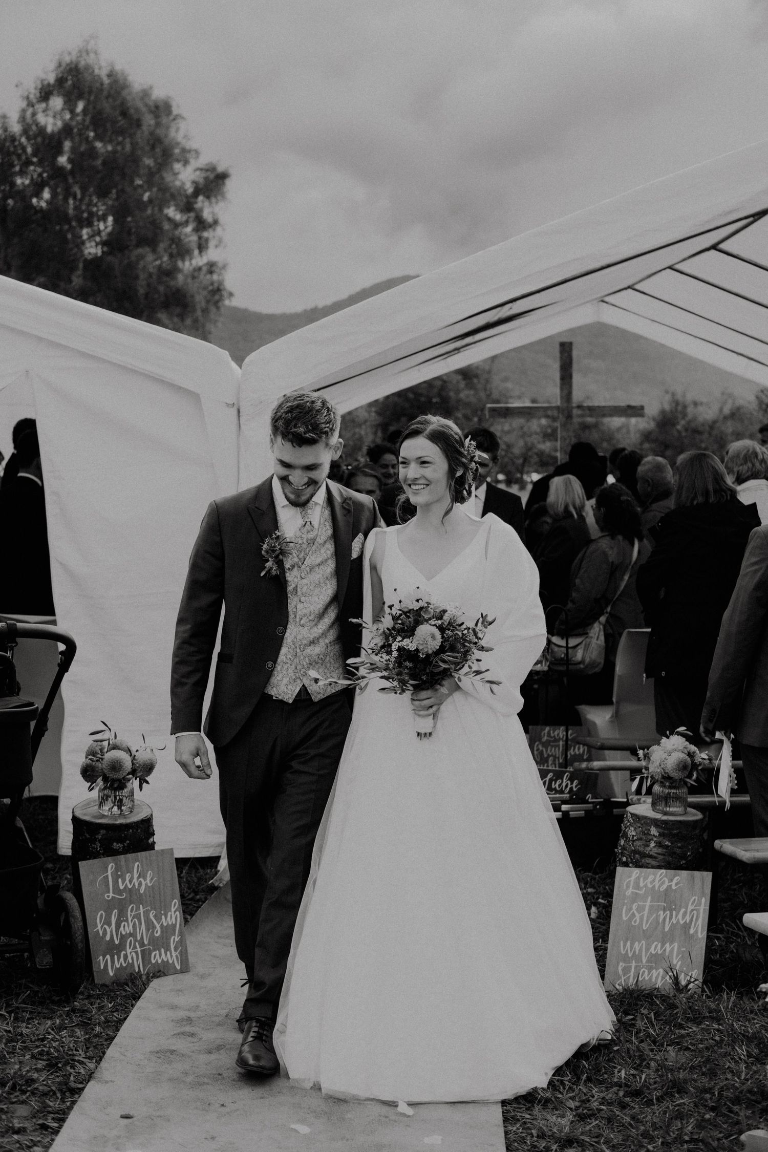 A couple in formal wedding attire walks together holding drinks at an outdoor reception with white tents.