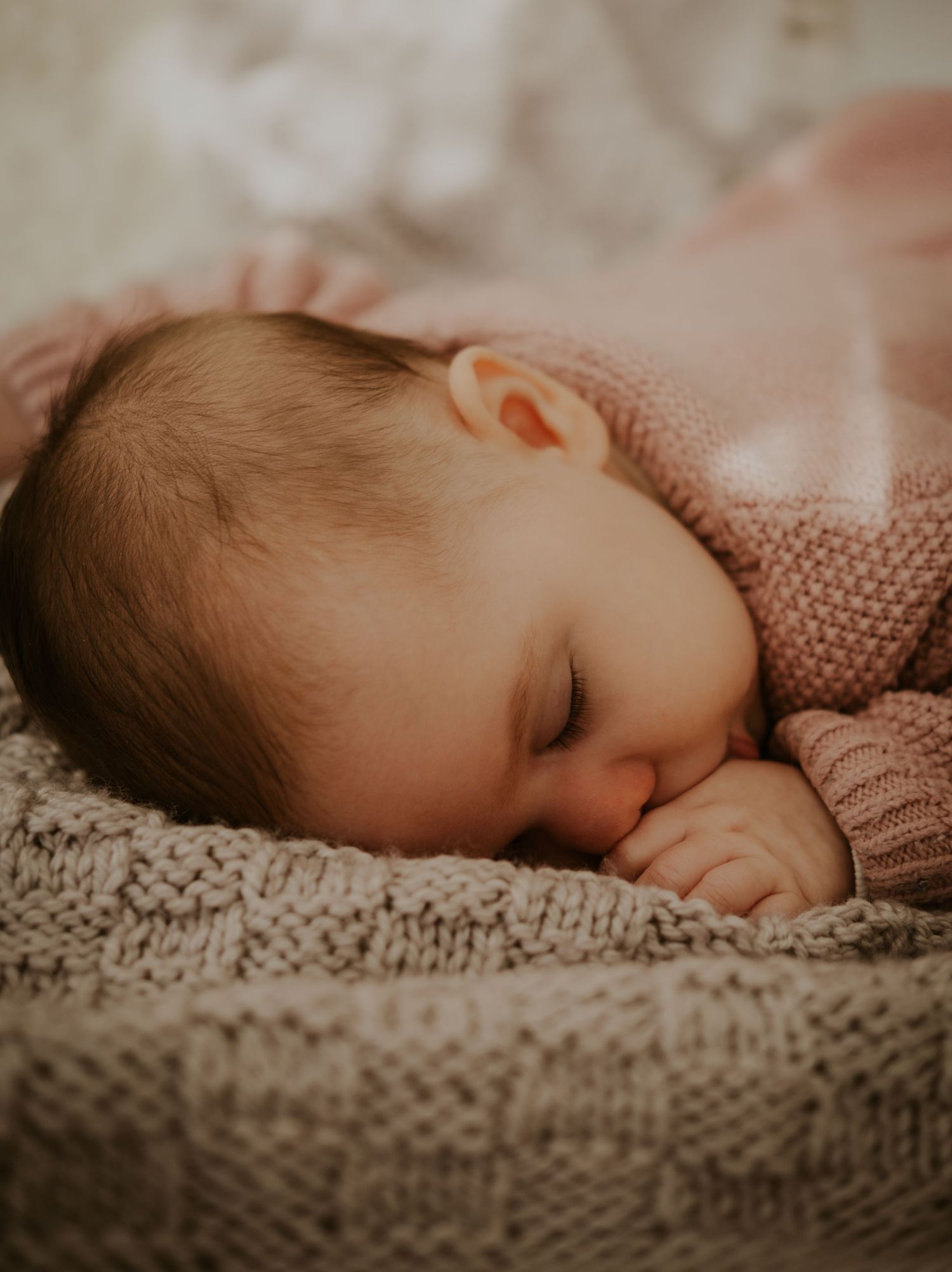 A sleeping baby in a pink sweater rests peacefully on a textured knit blanket in warm, muted lighting.