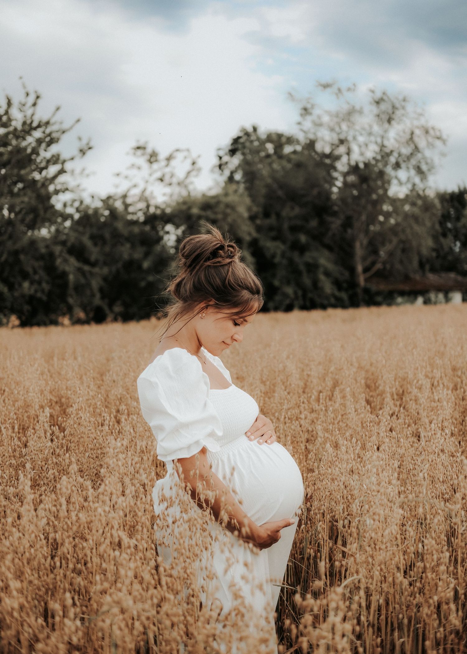 A person in a white dress stands in a golden wheat field on a cloudy day, creating a serene maternity portrait.