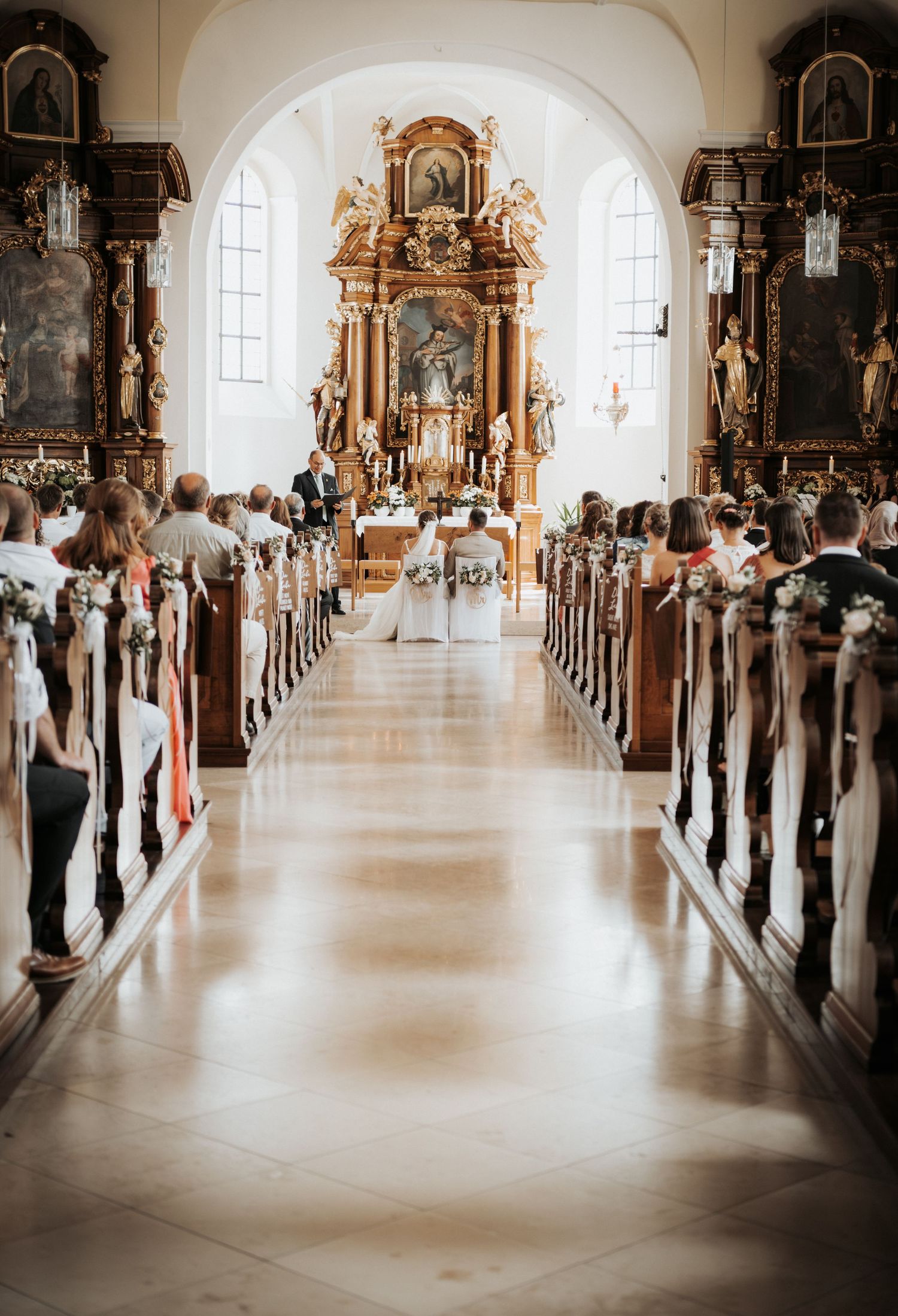 Elegant church wedding ceremony with white floral decorations along the pews leading to an ornate golden altar.