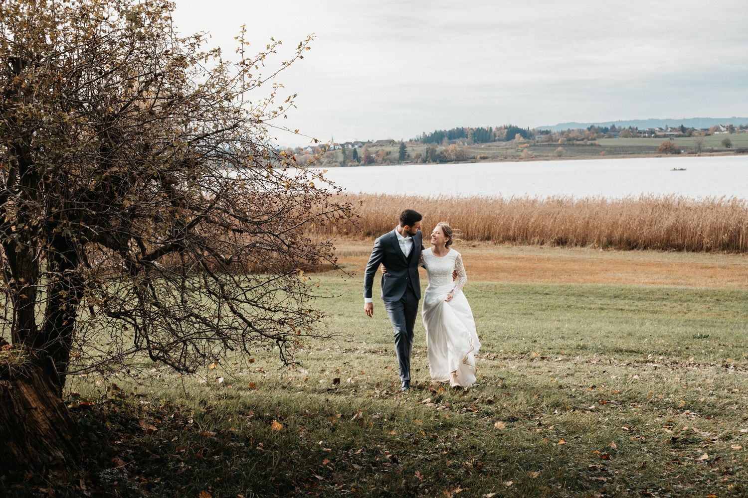 A couple in wedding attire walks through a scenic autumn landscape with a lake and rolling hills in the background.