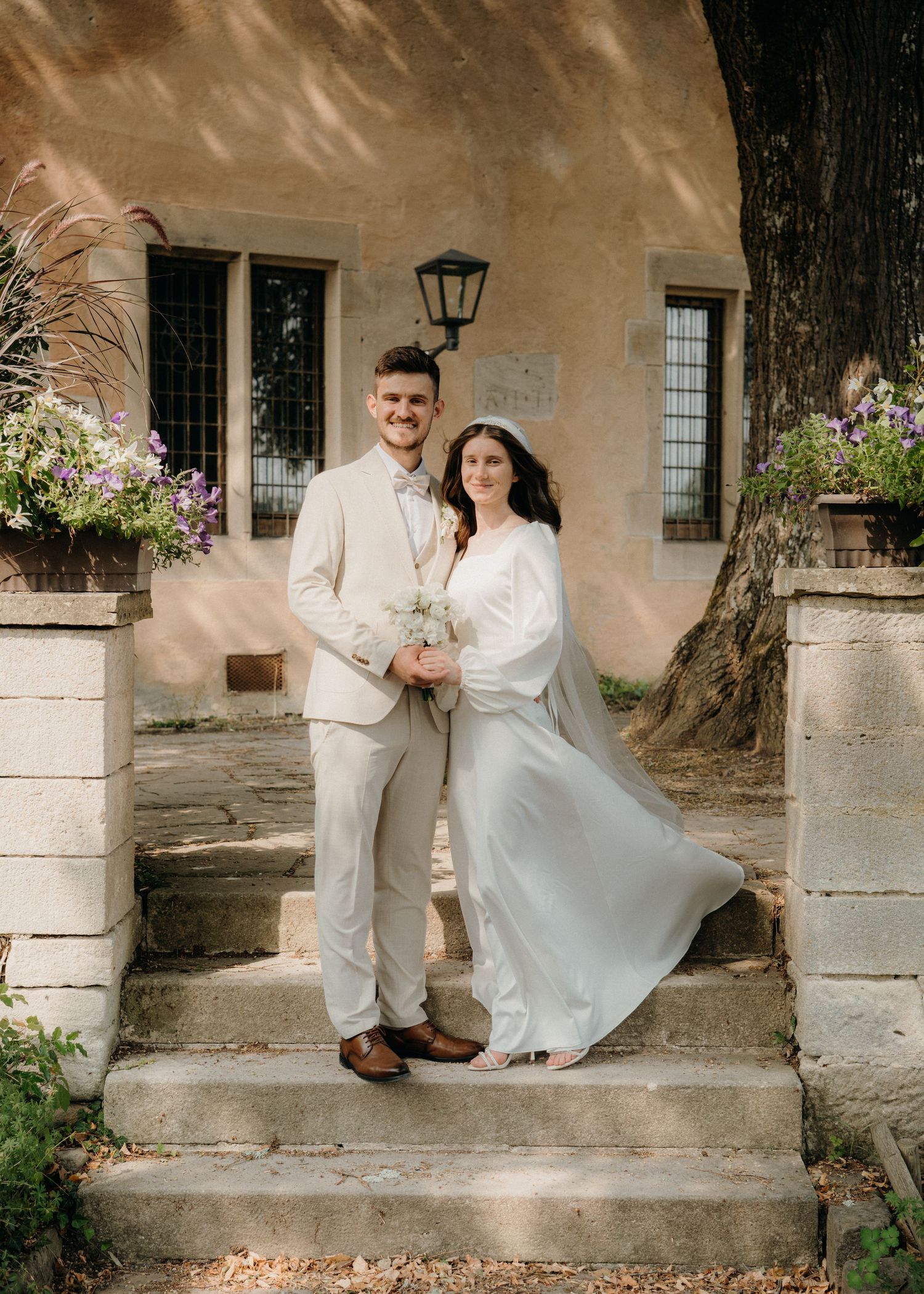 Bride and groom pose on stone steps outside a rustic venue, she holds white flowers as her gown flows in the breeze.
