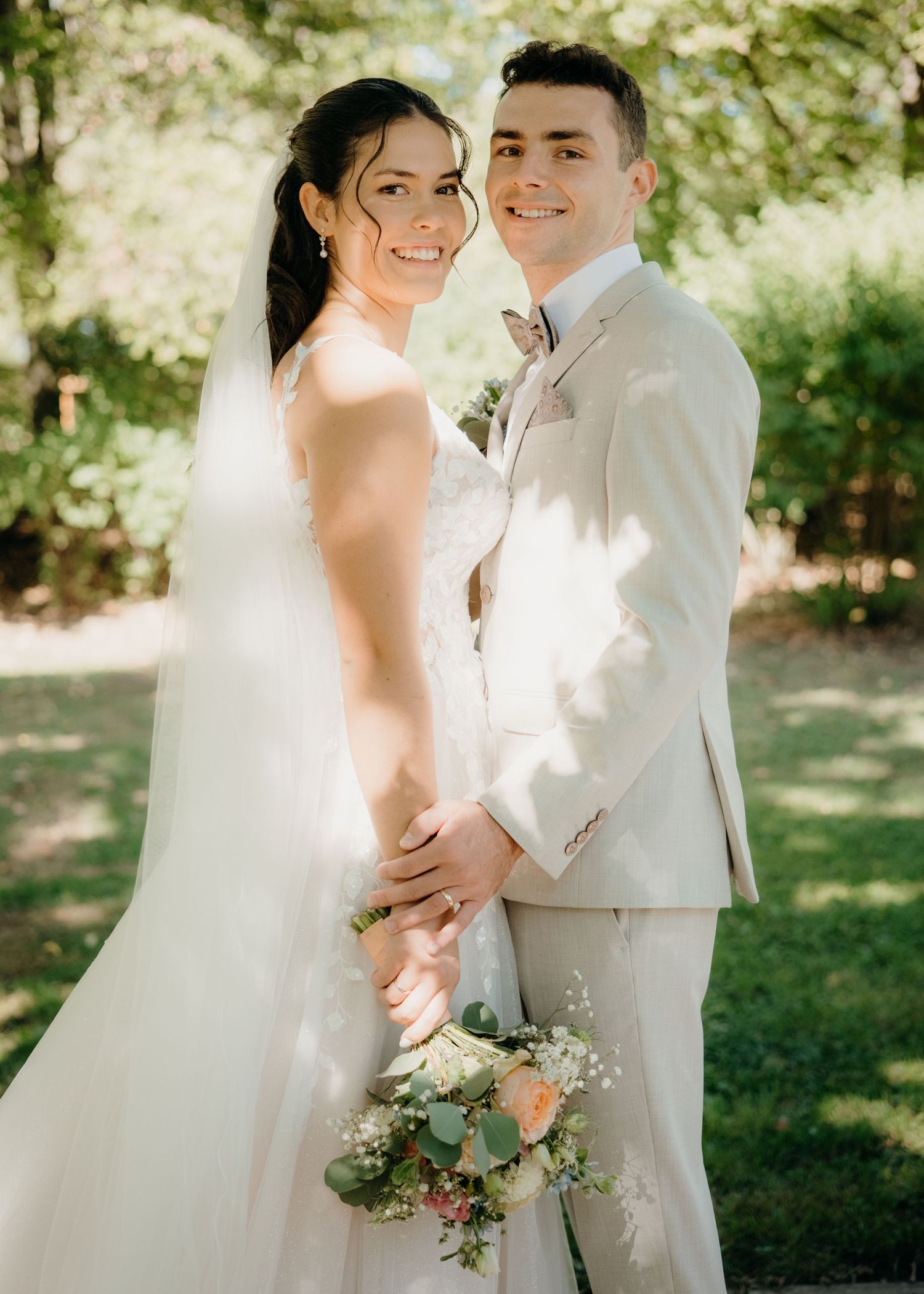 A smiling bride and groom pose together outdoors in sunlight, she holds a bouquet, he wears a beige suit.