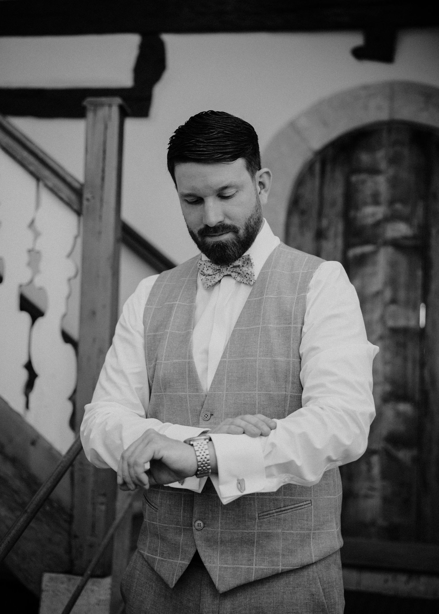 Groom in plaid vest and bow tie adjusting cufflinks outdoors in black and white wedding photo.