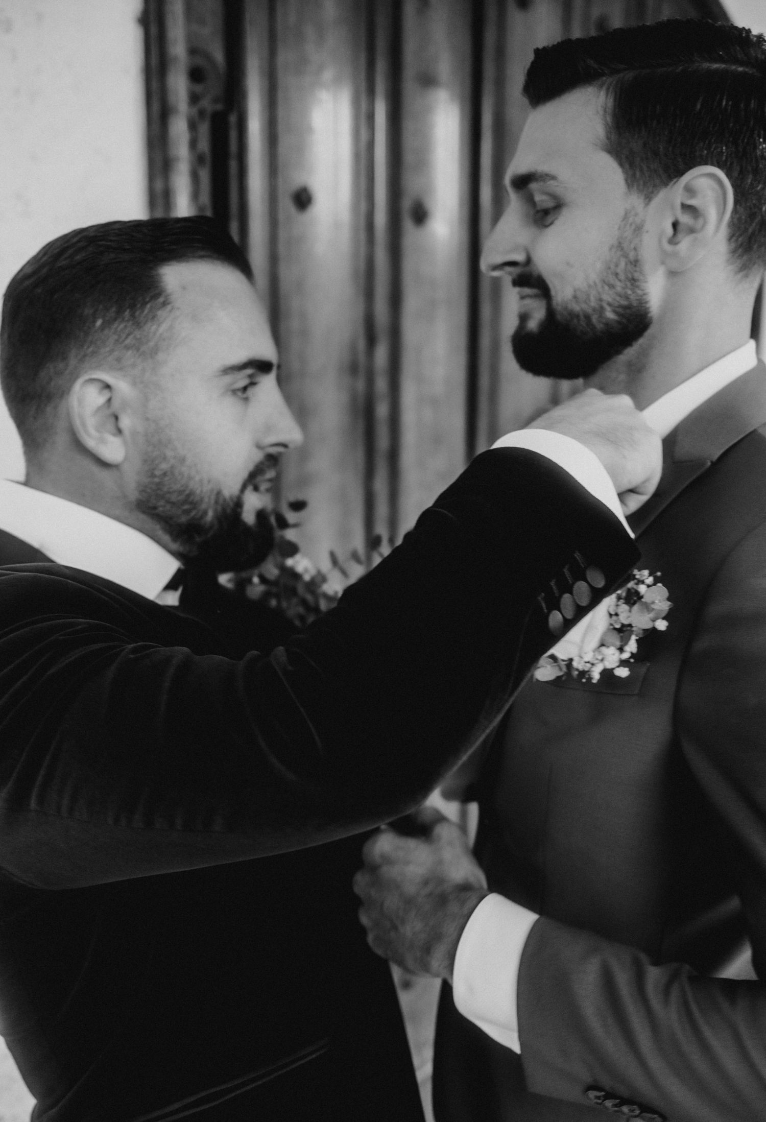 Black and white wedding photo showing two grooms in formal attire adjusting each other's boutonnieres.