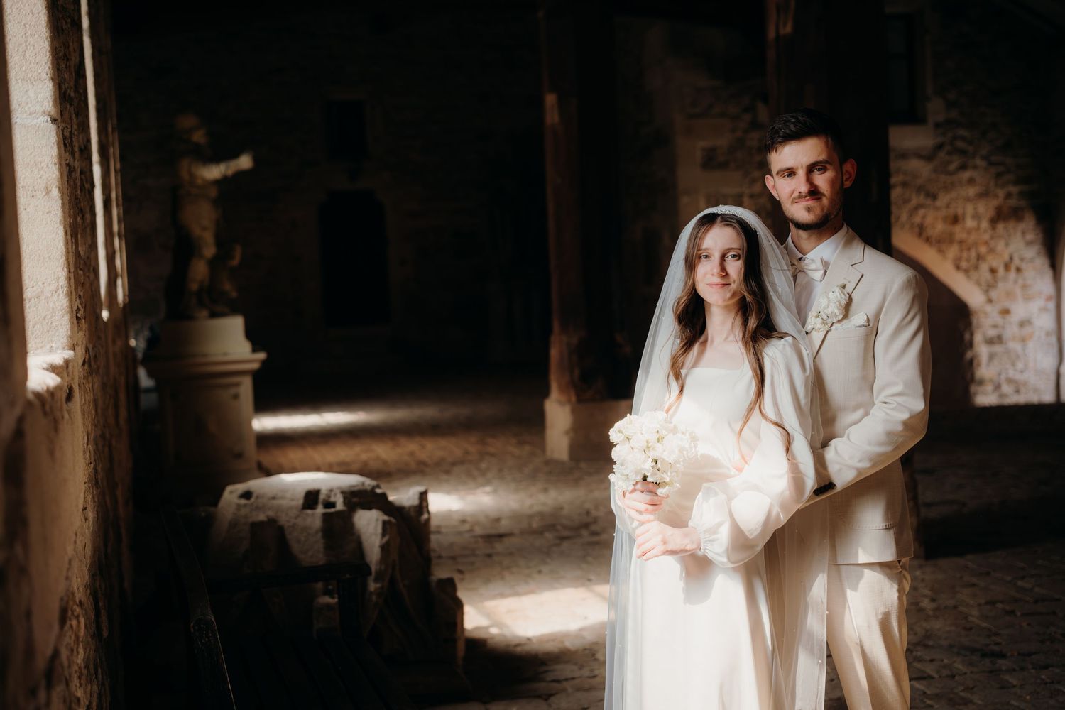 Bride and groom pose in rustic stone venue, she wears white gown with veil, he wears ivory suit.