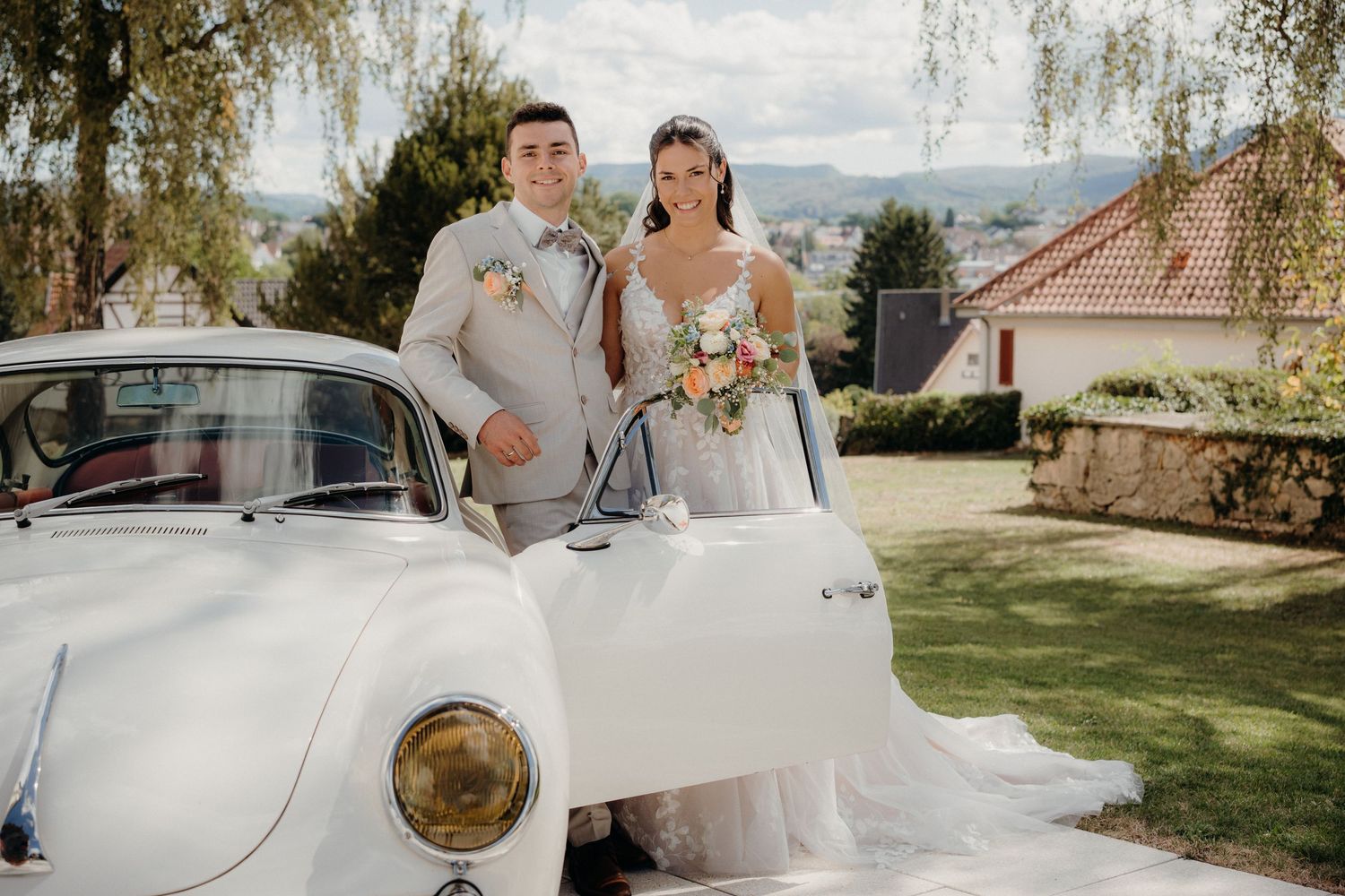 Bride and groom posing beside a vintage white car on their wedding day, surrounded by lush greenery.