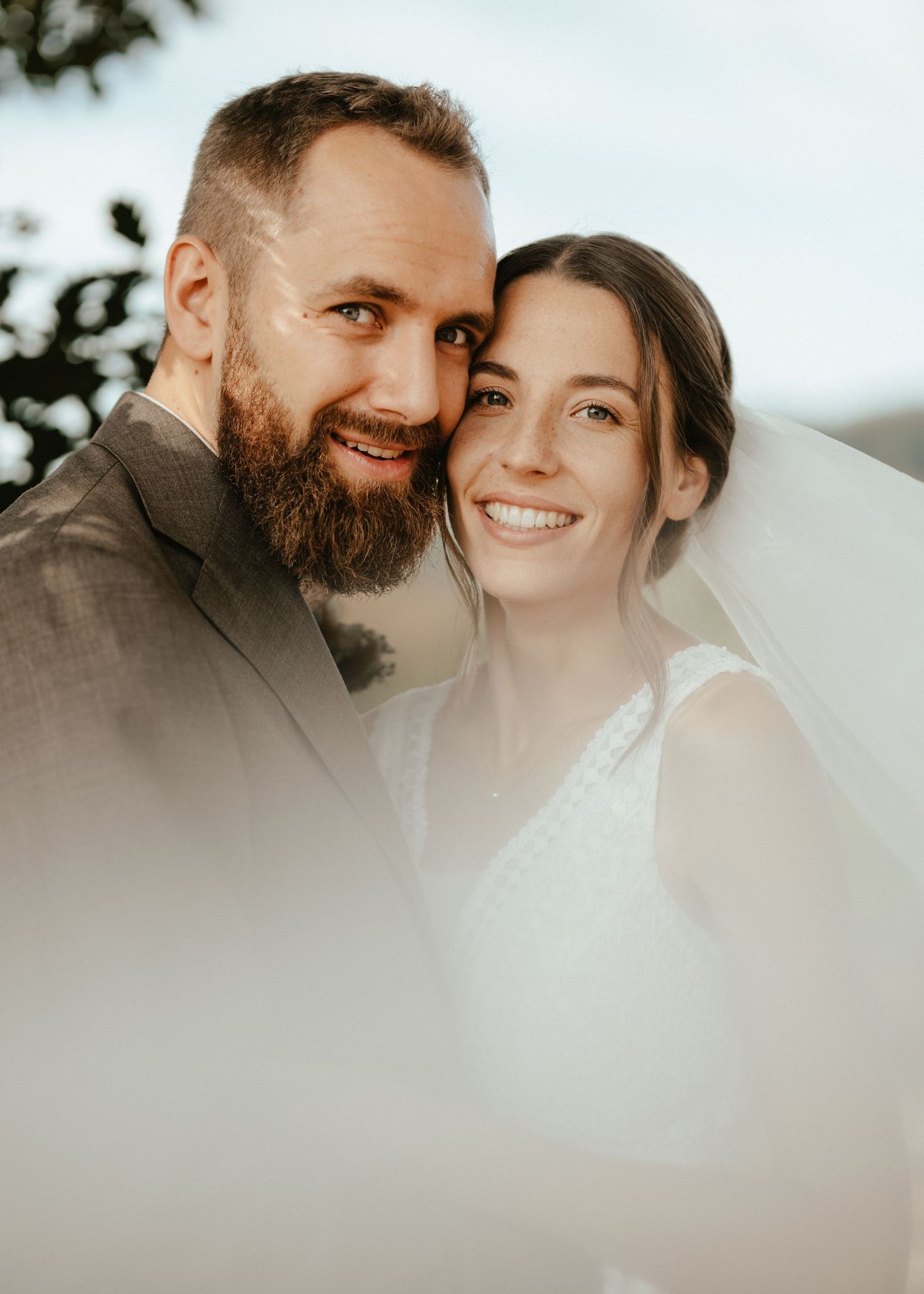 A bearded groom in a gray suit and his bride in a white veil smile closely together in a romantic outdoor wedding portrait.