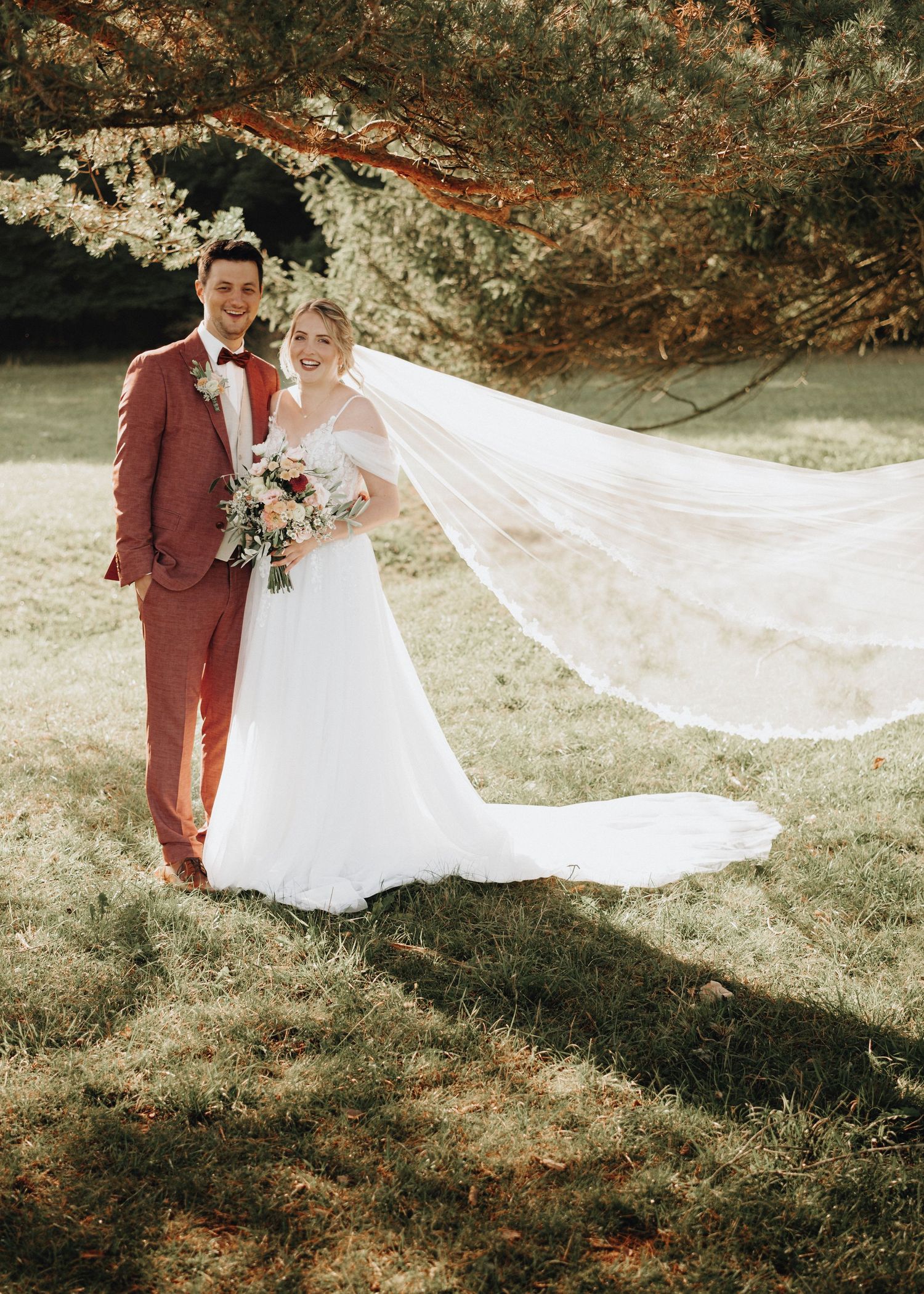 A couple in wedding attire poses outdoors with a flowing veil in autumn, the groom wearing a burgundy suit.