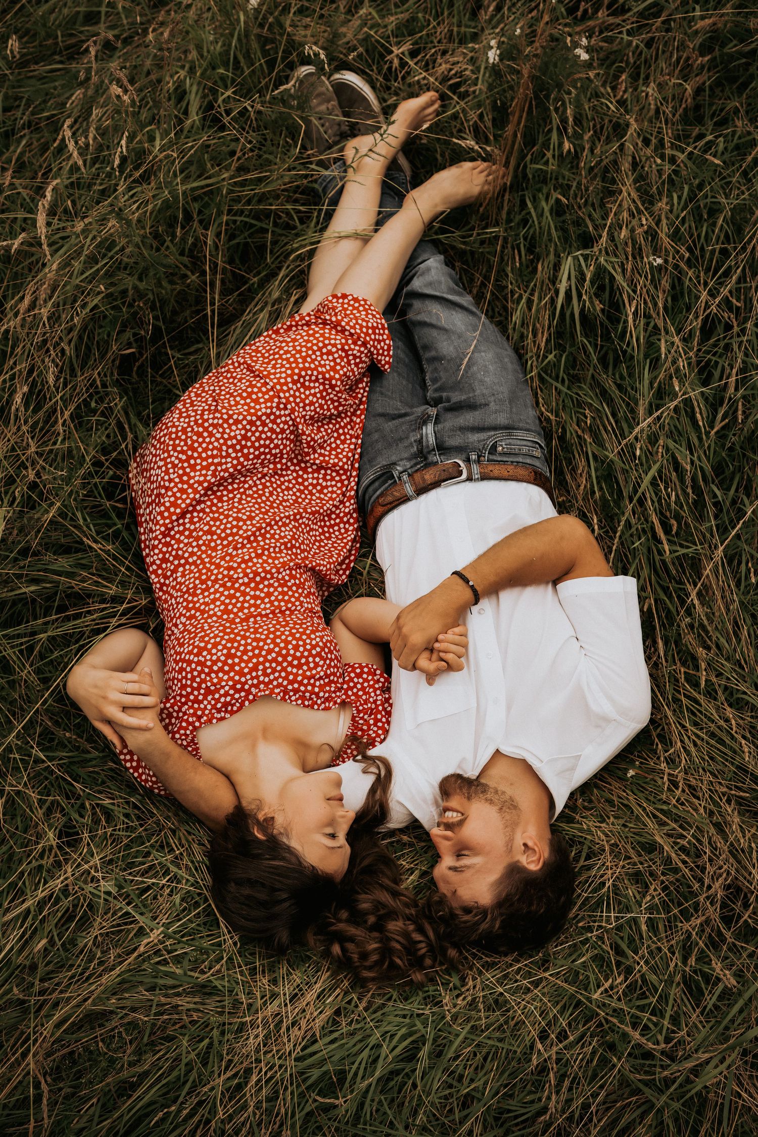 A couple lies together in tall grass wearing red polka dot dress and white shirt, captured from above in romantic poses.