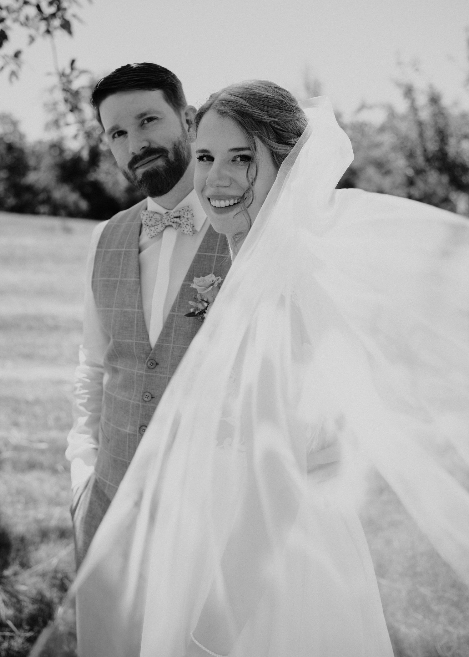 Black and white wedding photo of smiling bride with veil blowing and bearded groom in plaid suit outdoors.