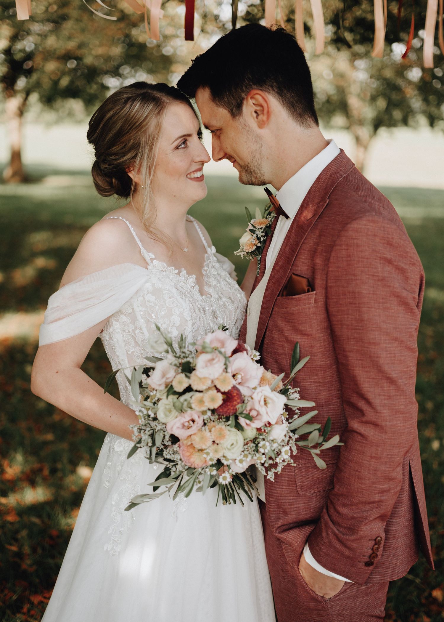 A couple in wedding attire share an intimate moment outdoors with the bride holding a pink and white floral bouquet.