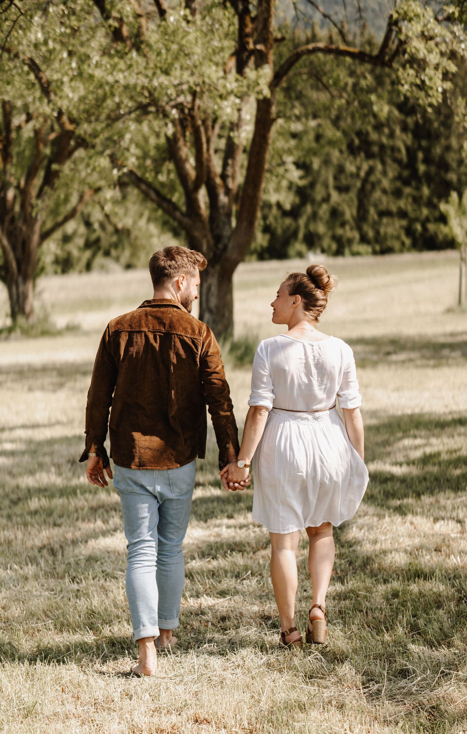A couple holding hands while walking through a sunny meadow wearing casual attire and brown leather jacket.