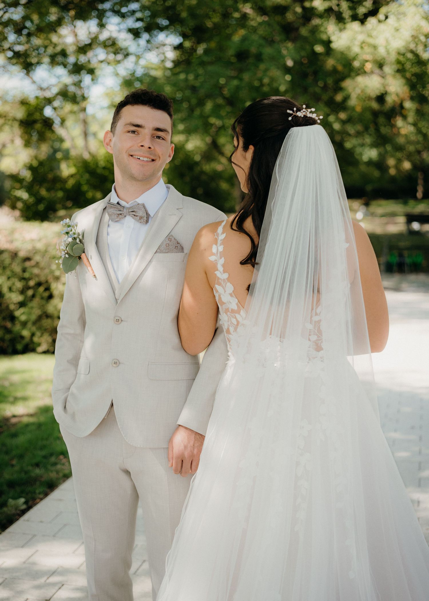 Bride in lace wedding gown with veil and groom in beige suit with bow tie posing outdoors on sunny day.