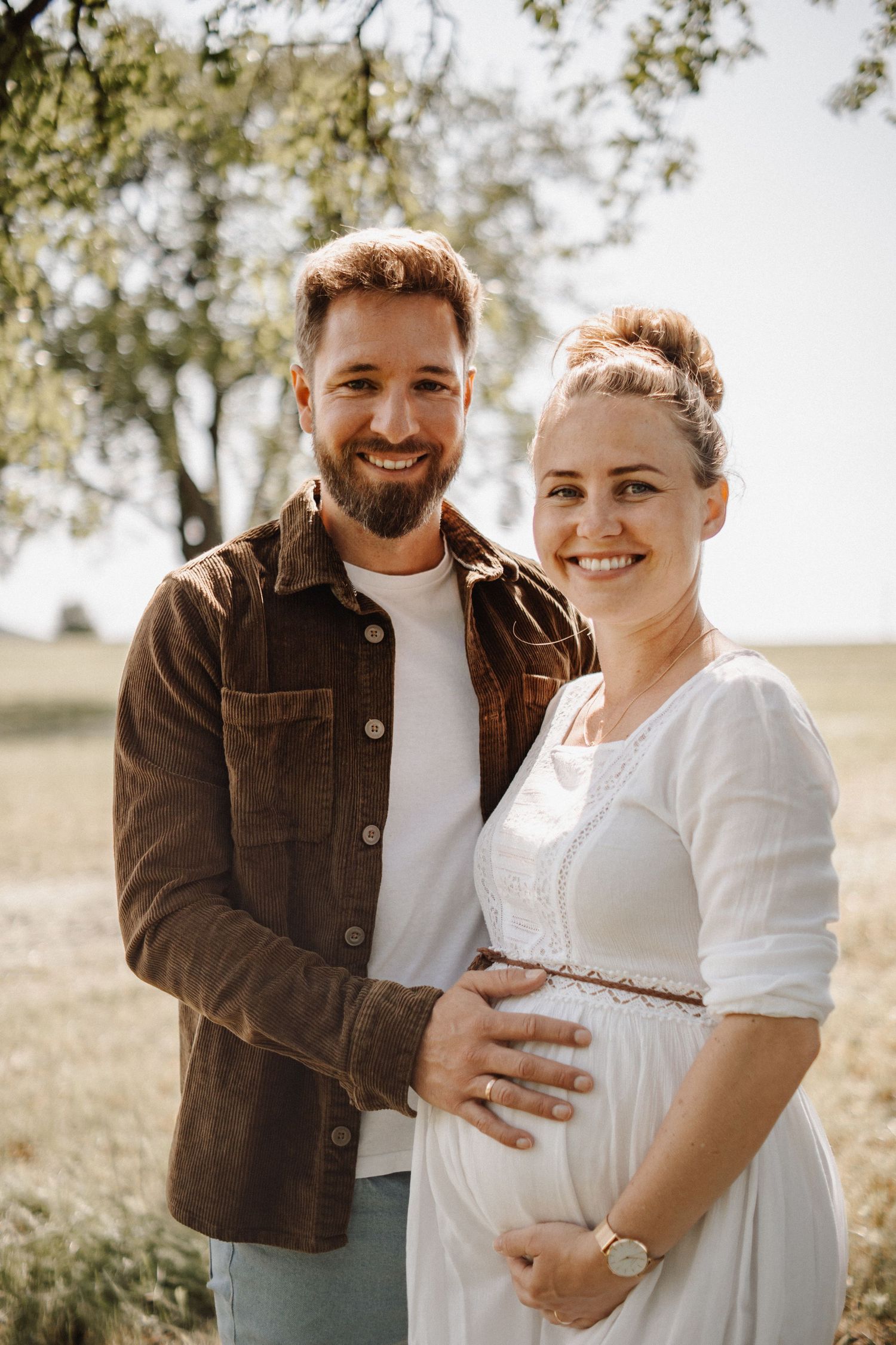 A couple in a brown jacket and white dress embrace in a sunny outdoor field setting during a maternity photoshoot.