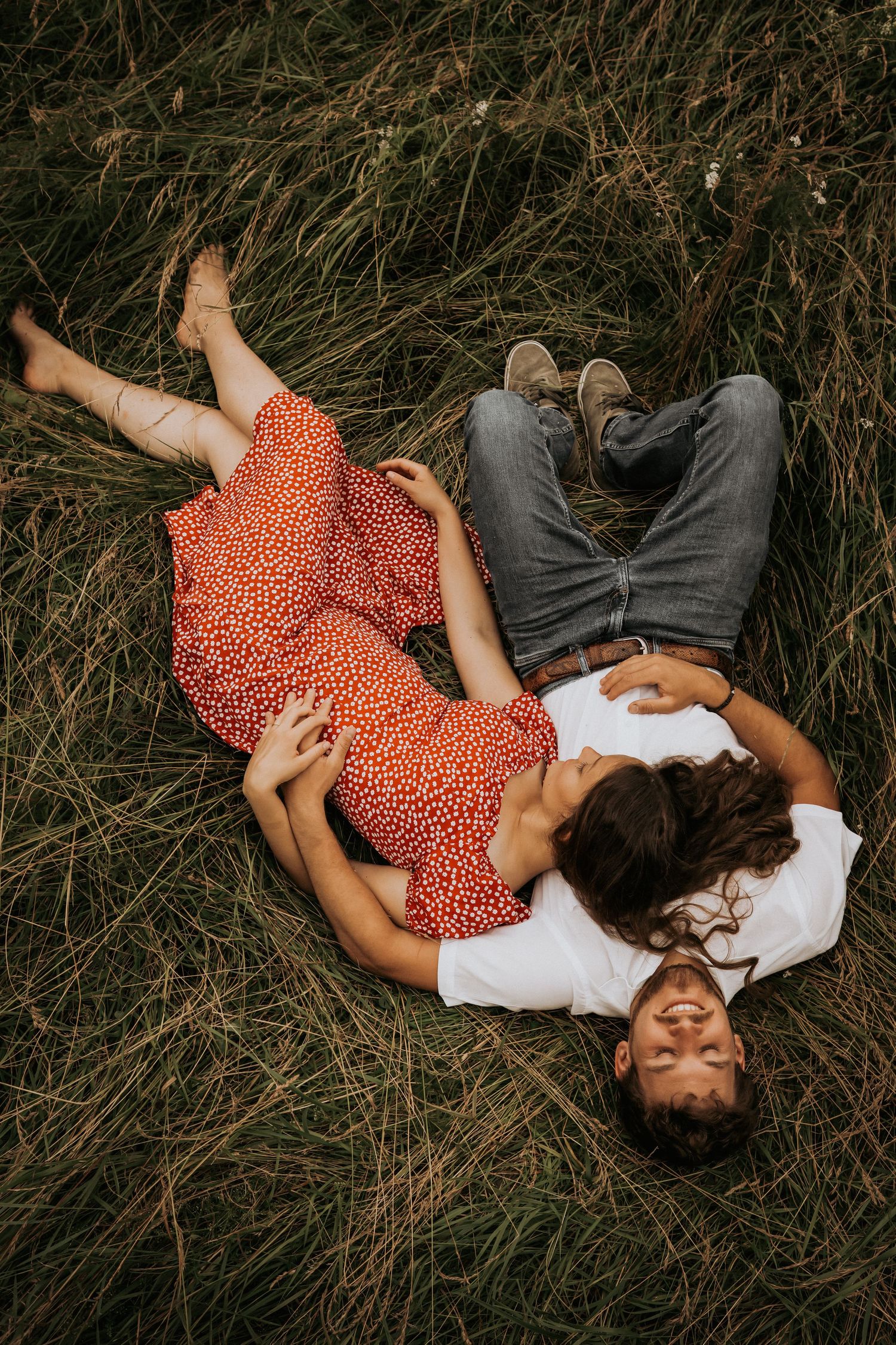 A couple in casual attire lies together on tall grass in a romantic outdoor photoshoot series.