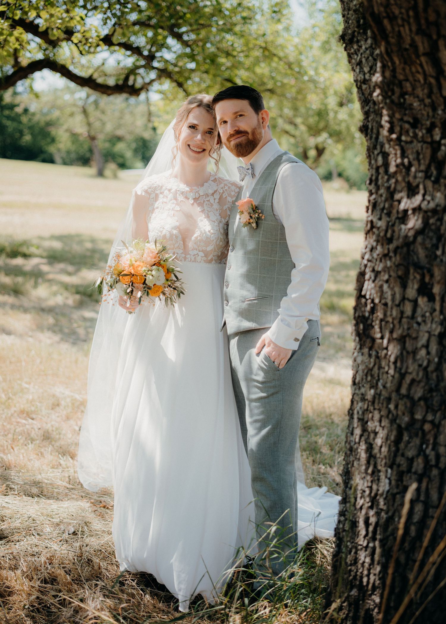 A bride and groom pose together near a tree outdoors, she holds an orange and white bouquet, he wears a gray vest.
