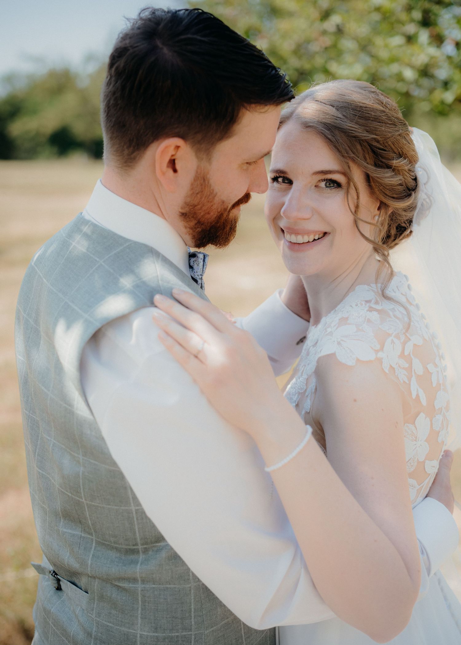 A smiling bride in lace wedding dress and veil embraces her bearded groom in a grey checked suit outdoors.