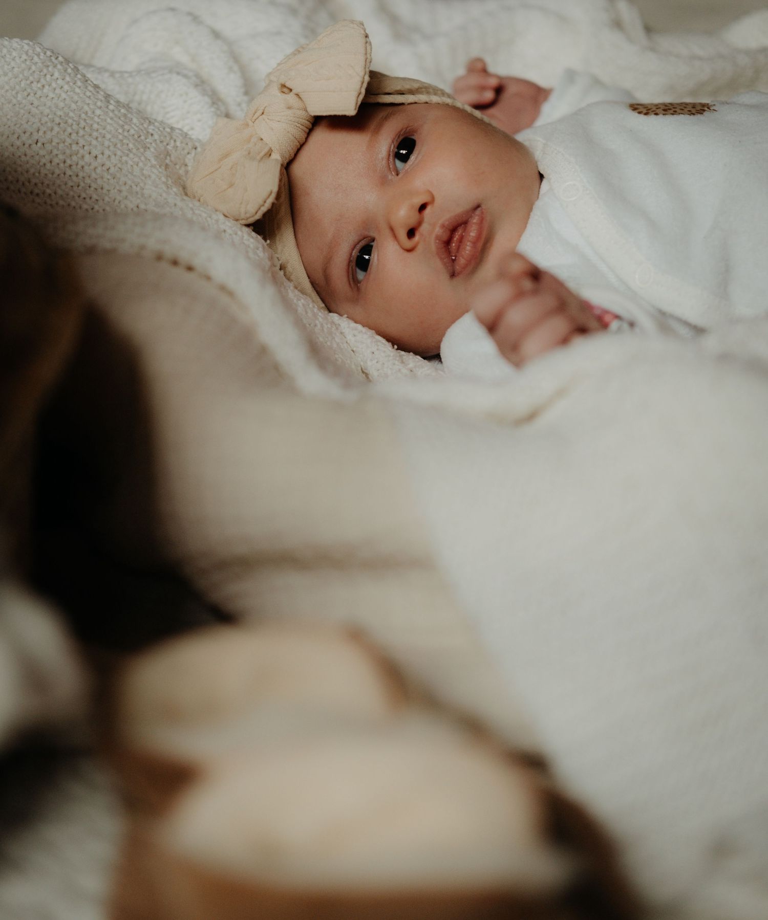 A baby in white clothing lies on soft blankets with a delicate bow accessory.