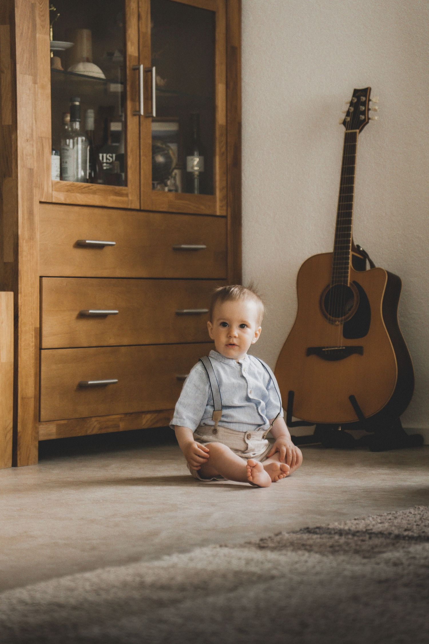 A small child sits on a carpeted floor in a room with wooden cabinets and a guitar visible in the background.